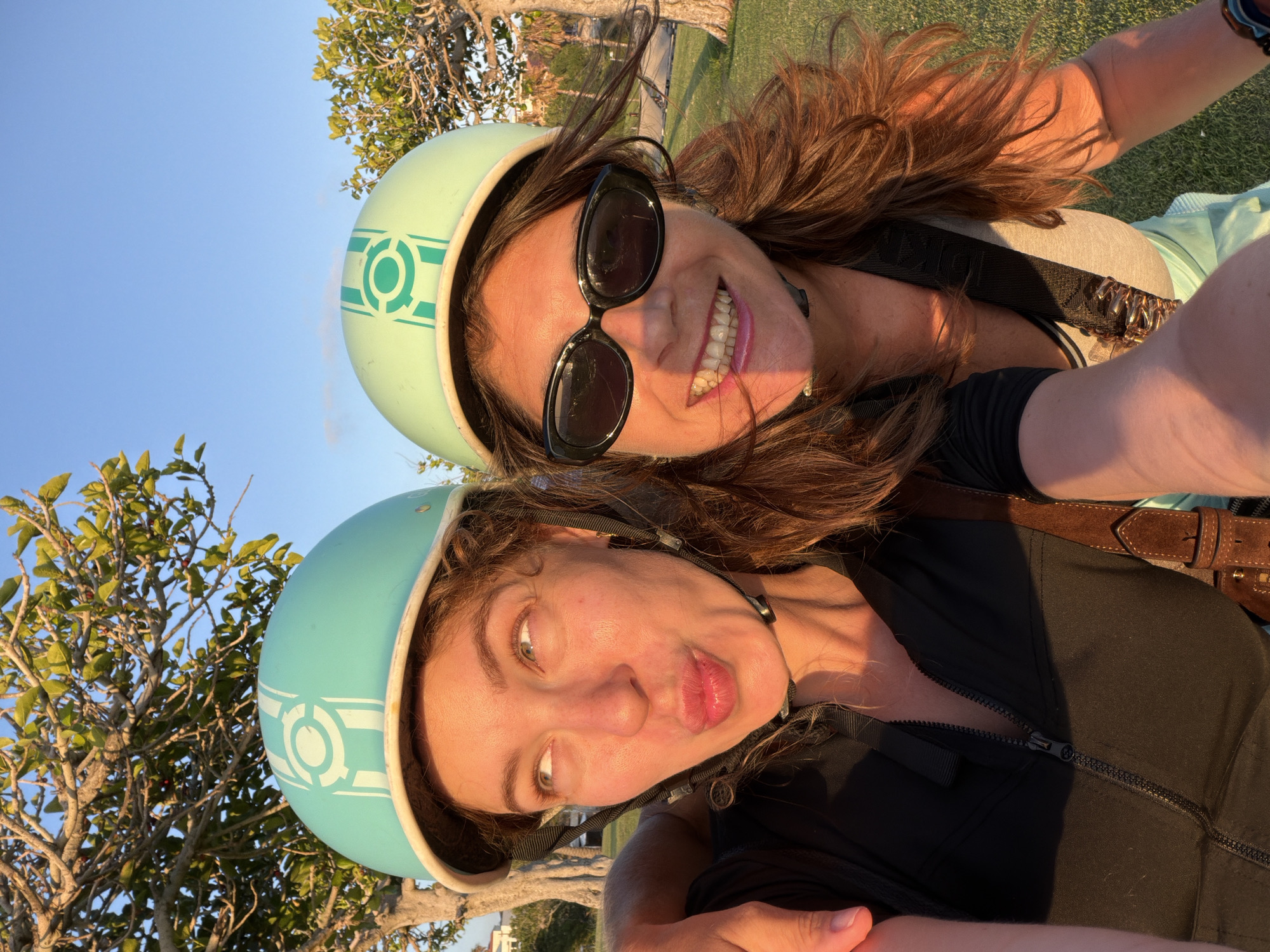 Two women are captured in a close-up, selfie-style photograph, posing outdoors with a clear blue sky and lush green grass as the backdrop. They are both wearing light blue helmets with a white circular emblem. The woman on the left has her hair down and is making a kissing face, while the woman on the right is smiling and wearing sunglasses. Sunlight casts a warm glow on their faces and hair. The setting appears to be a park or a grassy area, possibly in a suburban or coastal environment. There are trees in the background, and one of the women has her arm around the other's shoulders. The overall atmosphere of the image is cheerful and casual.