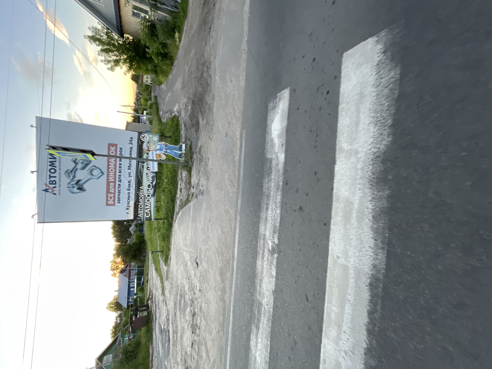 The image depicts a pedestrian crossing on a rural road in Krasnye Baki, Russia, with a billboard advertising an auto parts store and car wash in the background. The billboard, featuring Russian text that translates to "ALL FOR FOREIGN CARS" and "SPARE PARTS FOR ALL MAKES," is prominently displayed. Below the main advertisement, smaller text indicates "AUTO WASH SAMOOCHISTKA" and an address: "Krasnye Baki, ul. Michurina, 24a." A cutout of a cartoonish male figure, possibly representing a car wash employee, stands next to the billboard. The scene is set during dusk, with the warm glow of the setting sun visible in the sky. The road is asphalt, and the pedestrian crossing is marked by white stripes. Surrounding the road are grassy areas, trees, and residential buildings, characteristic of a village or small town. The overall impression is that of a quiet, everyday scene in a Russian settlement.