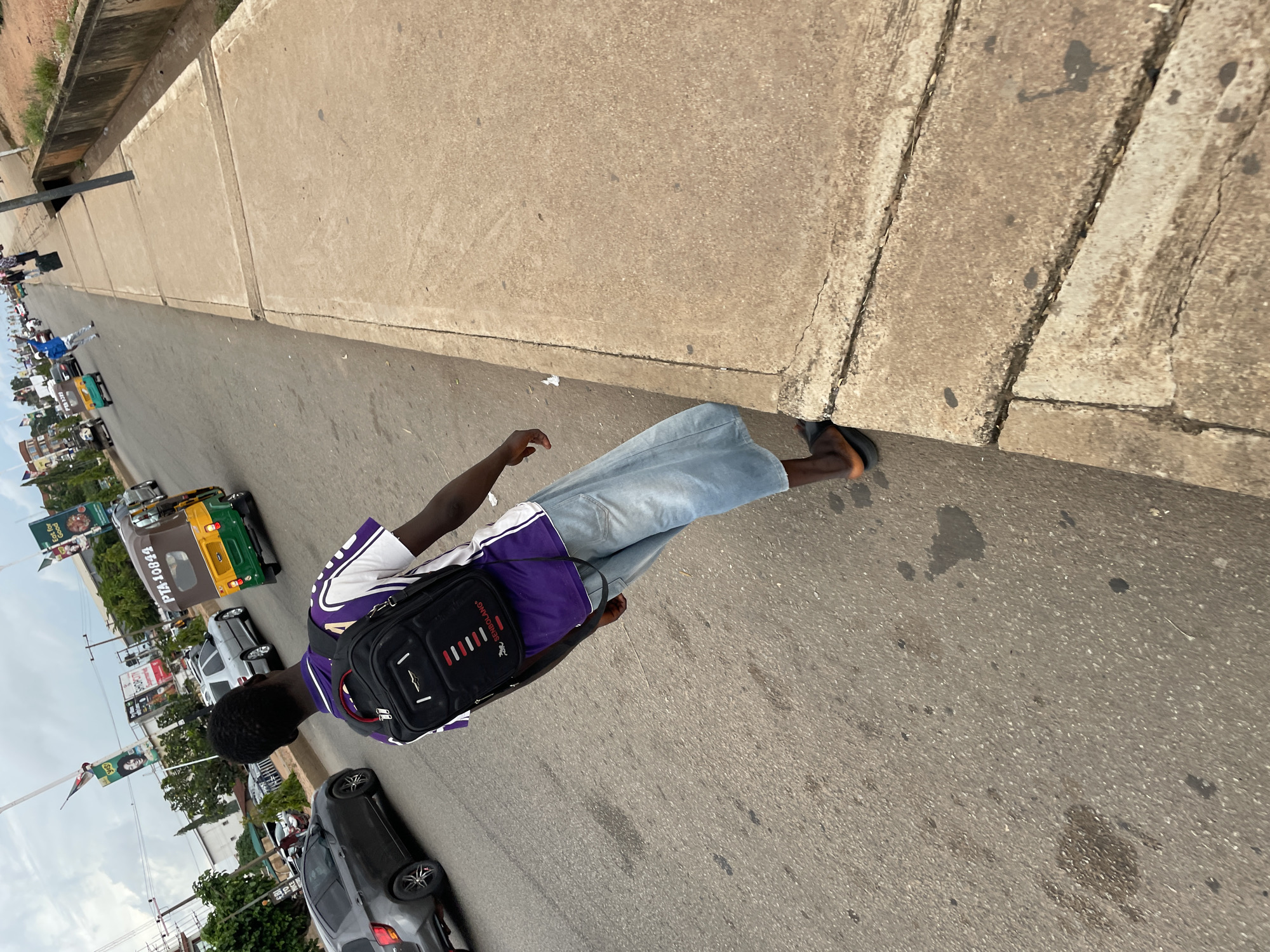 A young Black male is walking along the side of a busy road in Jos, Nigeria, on a cloudy day. He is wearing a purple and white t-shirt, light blue denim shorts, and dark sandals. He has a black backpack with red and white accents. The road is paved with asphalt and shows signs of wear and tear, including oil stains and cracks. To his right, there is a raised concrete sidewalk, which is also cracked and weathered. The background shows traffic, including cars, a yellow and green tricycle taxi, and some buildings. A man in a blue shirt can be seen in the distance, walking on the sidewalk and raising his arm. The scene suggests a typical urban environment with ongoing activity.