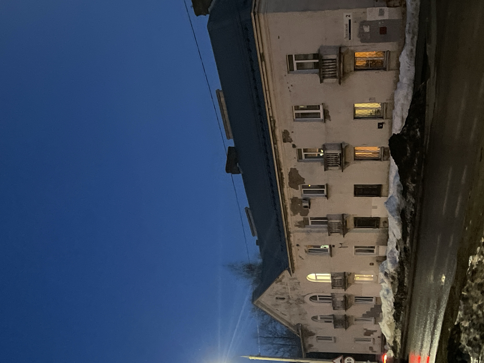The image shows a residential building in Izhevsk, Russia at dusk. The building is several stories tall with windows and balconies. Some windows are lit. Patches of snow are along the building's edge. The sky is a deep blue. A road runs alongside the building.