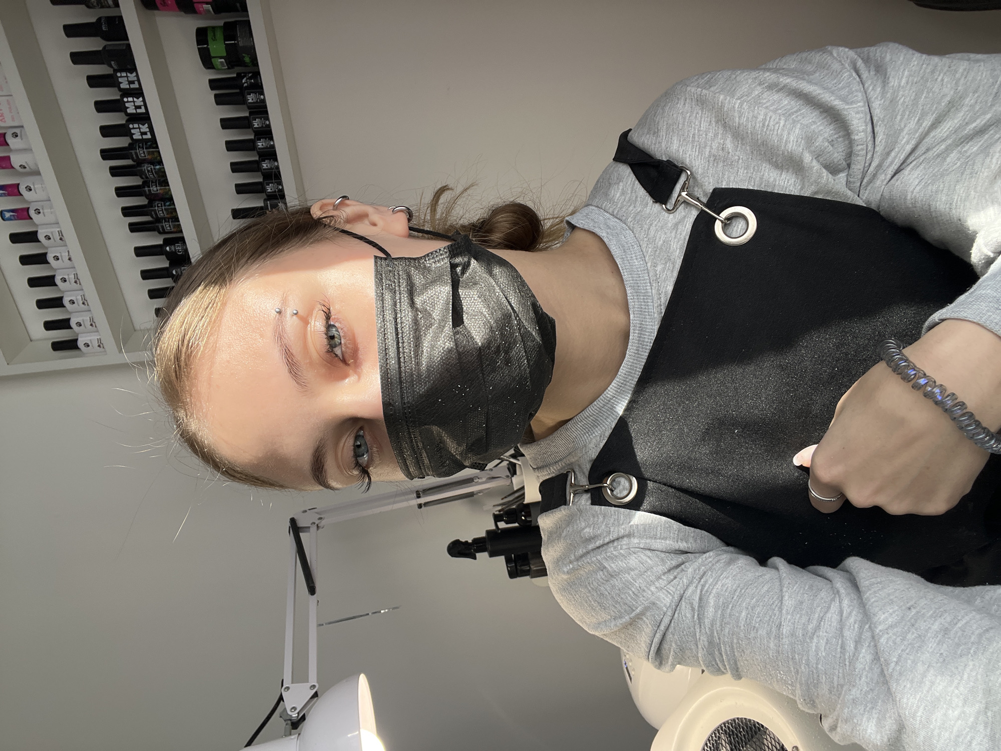 A young woman is prominently featured in a close-up shot, positioned vertically. She is indoors, likely in a nail salon or beauty studio in Фоки, Russia, indicated by the shelves of nail polish bottles visible in the background and specialized equipment in the foreground.
The woman has light brown hair, pulled back into a bun visible behind her ear. She wears a black disposable face mask covering her nose and mouth. Her visible blue eyes are open, and she has long eyelashes. An eyebrow piercing is visible above her left eye, and several silver hoop earrings adorn her left ear. She is dressed in a light gray long-sleeve shirt under a black apron-style top, secured with silver clasps and grommets. A bracelet made of dark, iridescent beads is on her right wrist, and a simple silver ring is on her right ring finger. Her left hand is partially visible, with a light-colored fingernail.
The background on the left features white shelves stacked with numerous small, dark bottles, presumably nail polish or gel. Visible text on some bottles includes "MIMI", "LK", and "GEL". On the right, part of a white professional desk lamp with an adjustable arm is seen, casting bright light. Below this, a white circular object with a grate, possibly a nail dust collector or fan, is partially visible. The scene is brightly lit, suggesting daytime, with strong natural light illuminating the woman's right side. The woman appears to be looking towards the camera, possibly posing or taking a selfie, and seems to be a professional working in the studio.
