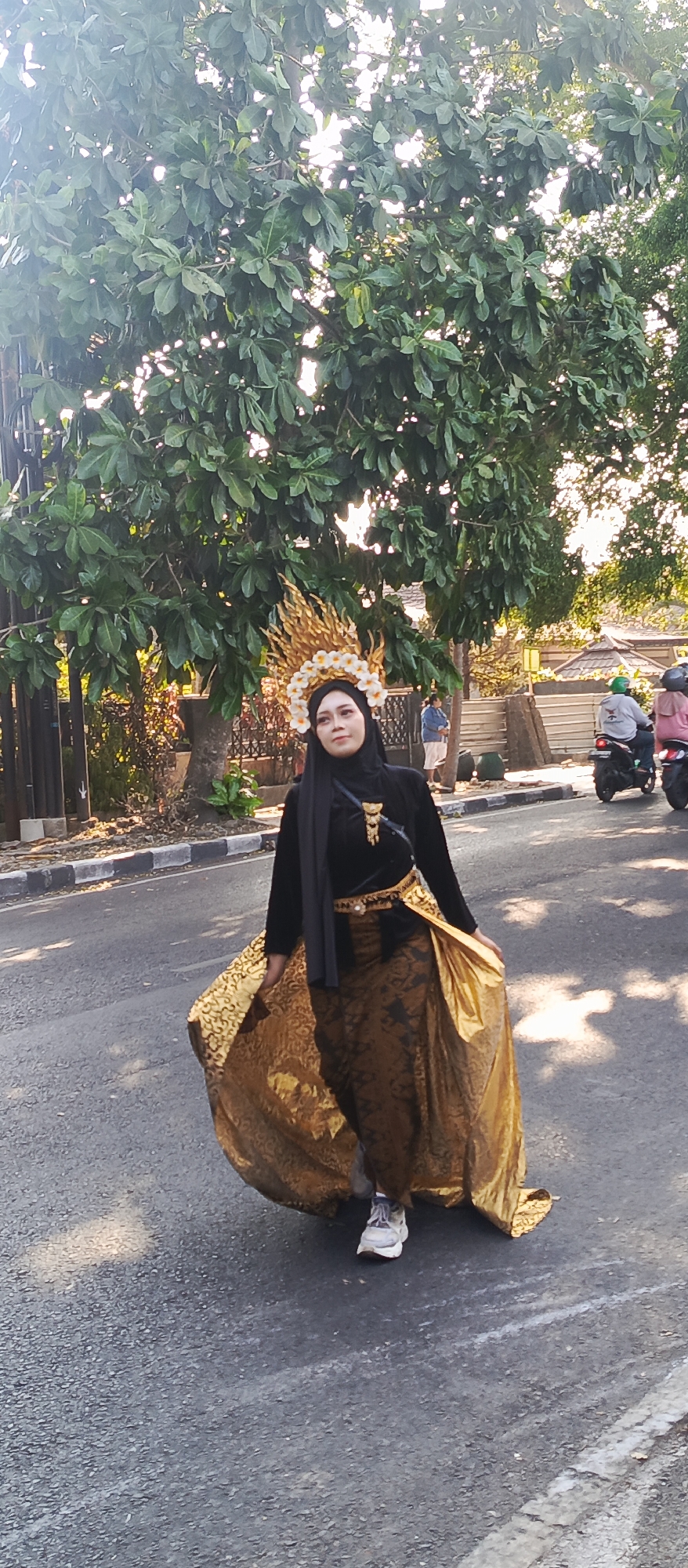 In Malang, Indonesia, a woman dressed in a traditional black Islamic hijab and a flowing golden brocade skirt with a matching sash walks on an asphalt road. She wears a striking golden headdress adorned with white flowers. Her outfit is completed with white sneakers. In the background, trees with green leaves line the street, and a few motorbikes are visible, with two parked on the right side of the road, one ridden by individuals wearing helmets and the other by a person with a green helmet. The scene appears to be outdoors, under daylight, with sunlight filtering through the trees, casting shadows on the road.