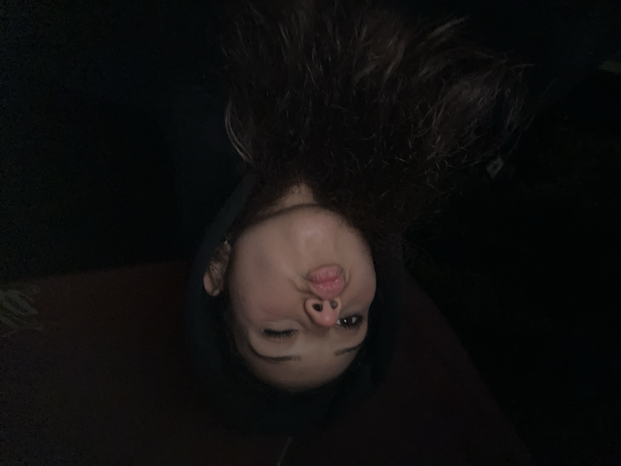 The image shows a close-up, low-light selfie of a young woman wearing a black hoodie. Her head is tilted, and she is winking and puckering her lips. Her long, dark, curly hair is visible, framing her face and falling down her shoulders. The background is dark and indistinct, with some muted reddish-brown tones visible at the top. There is no discernible evidence to suggest this image was taken in Zlatograd, Bulgaria, or any specific city.