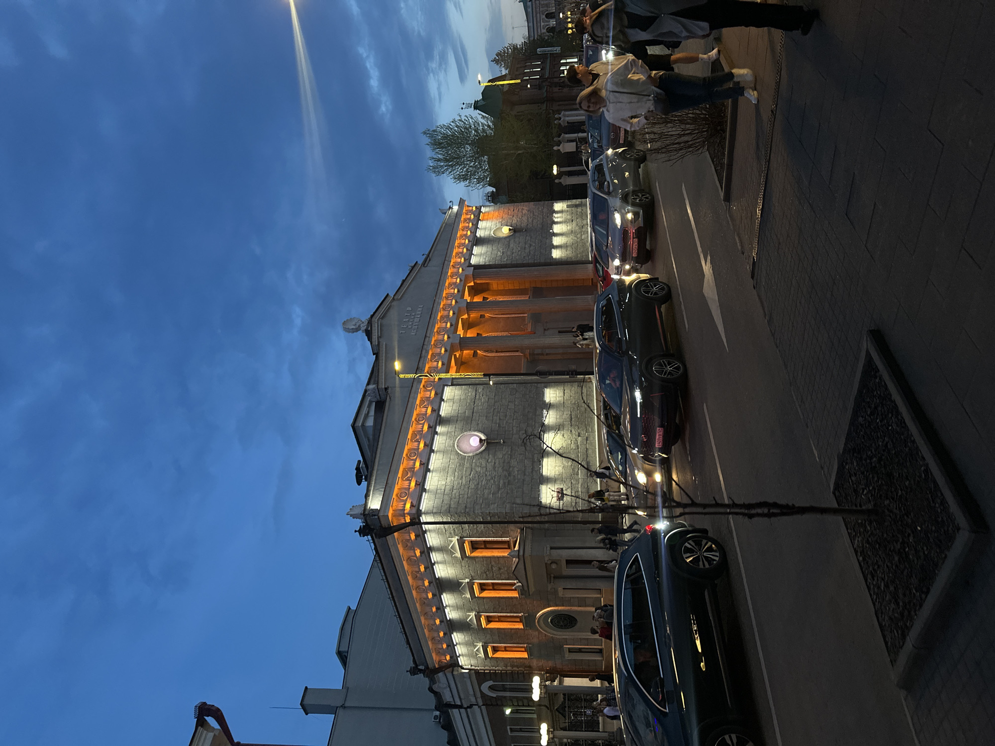 Captured at dusk, the image depicts the A.S. Pushkin Drama Theater in Krasnoyarsk, Russia. The three-story theater is prominently featured, with its facade illuminated by warm, orange lights, contrasting against the darkening blue sky. Architectural details such as columns and decorative molding are visible. Several parked cars line the street adjacent to the theater, with pedestrians walking along the sidewalk. A streetlamp shines brightly overhead. Other buildings and trees are visible in the background.