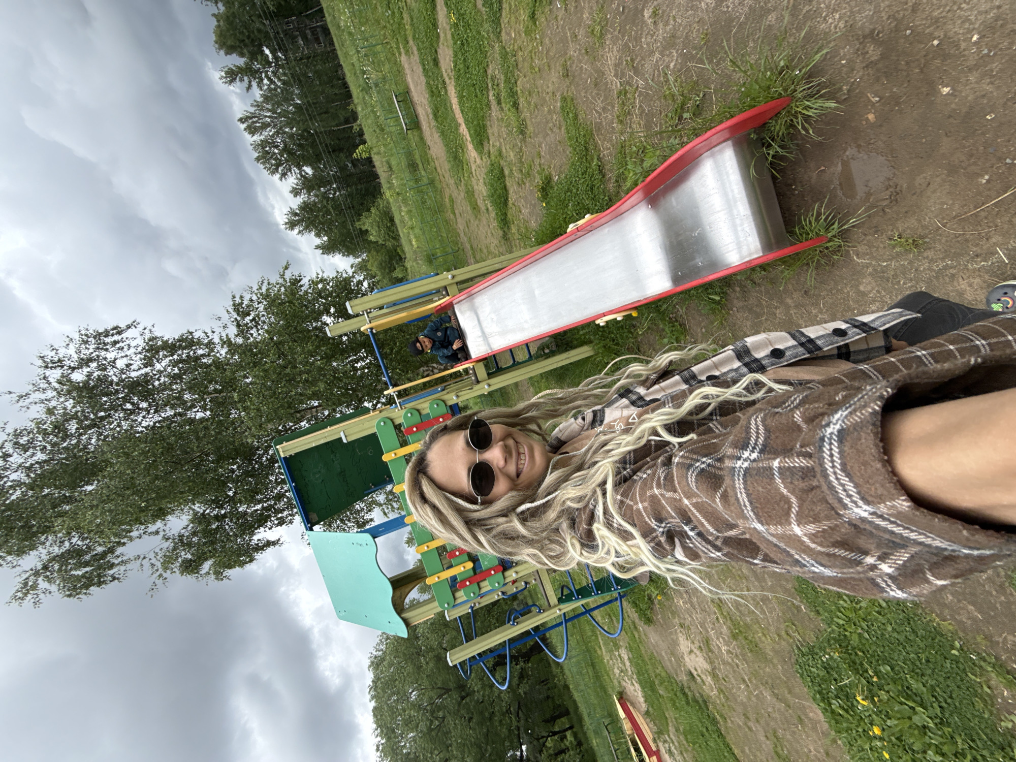 A woman with long, blonde dreadlocks and round sunglasses is taking a selfie in Rusanovo, Russia. She is wearing a brown plaid shirt. Behind her is a playground structure with a slide, climbing apparatus, and platform. A man is visible near the slide. The environment is a grassy area with trees, and the sky is overcast.
