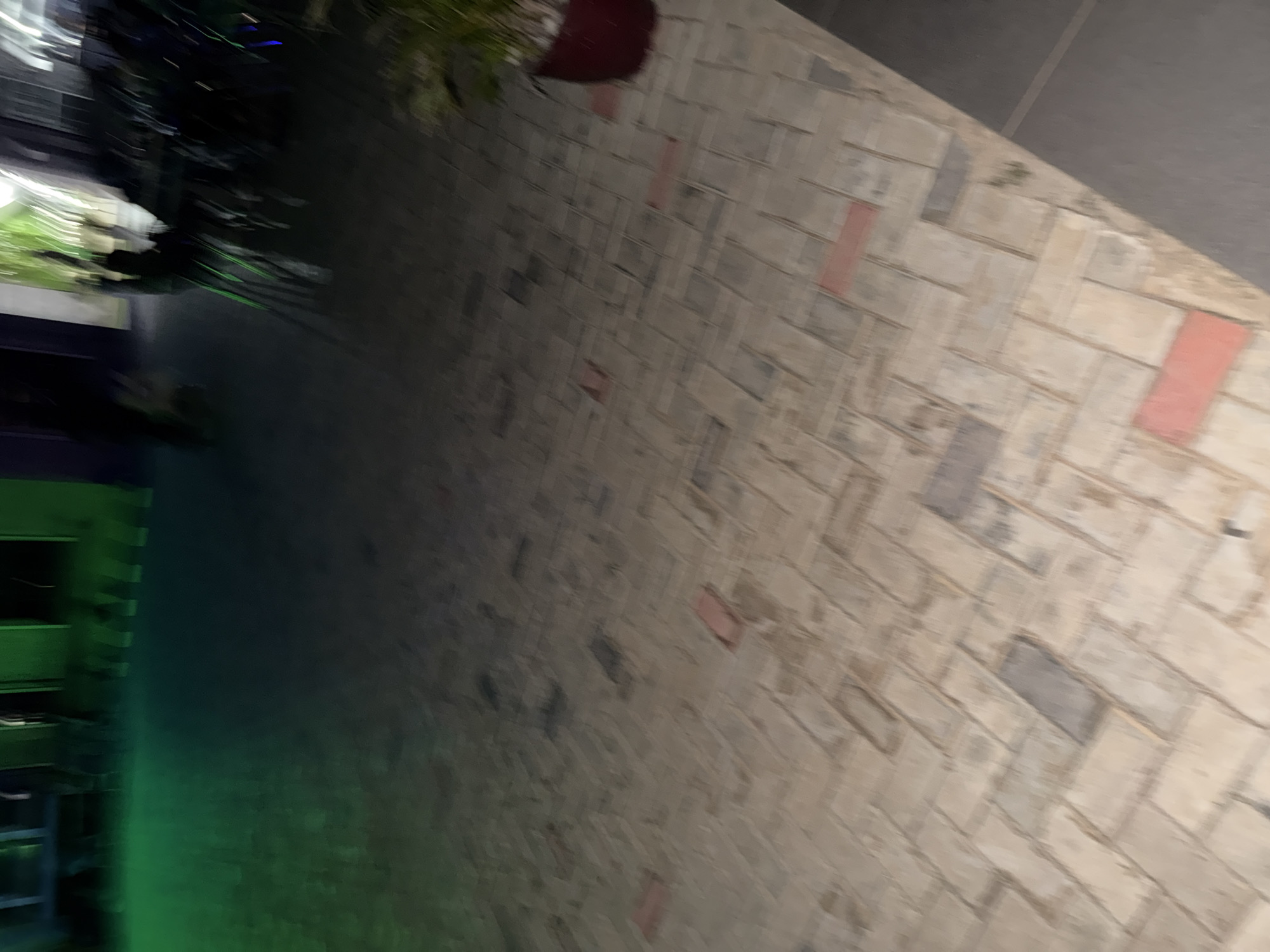 The image depicts a ground-level view of an outdoor tiled area. The tiles are laid in a herringbone pattern, with a mix of grey and reddish-brown rectangular stones. Some of the tiles appear wet or stained. In the background, there is a dark, blurry area with hints of green lighting, suggesting an indoor or covered space. To the far right, a dark red pot containing a green plant is visible. The overall impression is that of an exterior walkway or patio, possibly at a commercial establishment or residence. The location is identified as Bukuru, Nigeria.