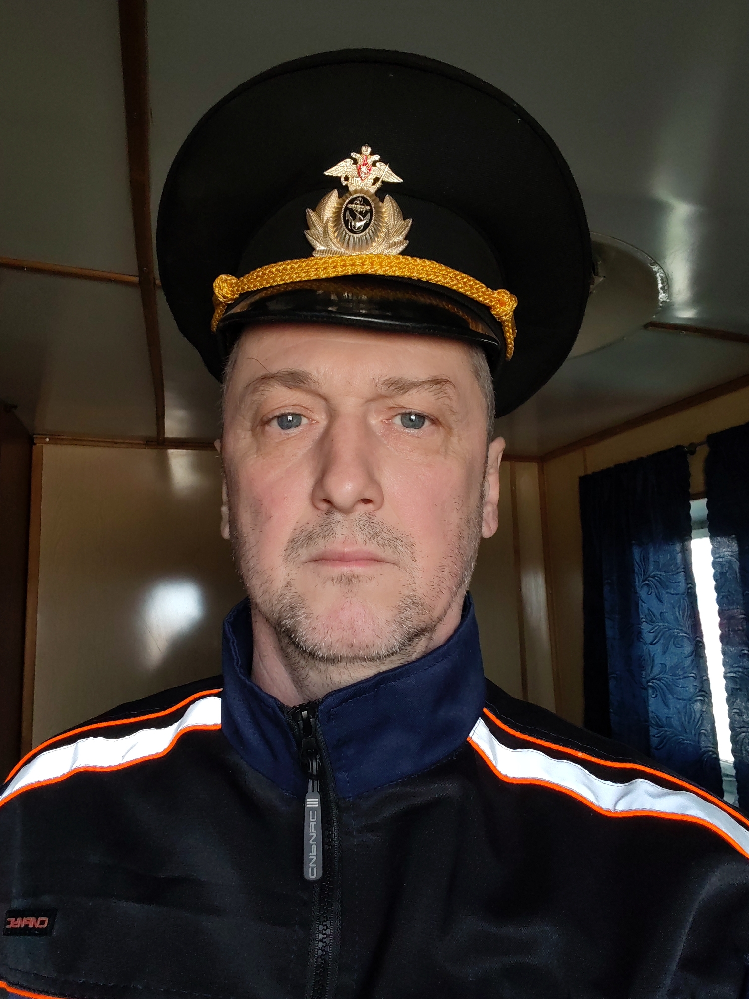 A middle-aged man with light blue eyes and short, light stubble is shown in a medium close-up shot. He is wearing a black peaked cap adorned with a gold emblem and a gold-colored twisted cord on the brim. He is also wearing a black jacket with a blue collar and shoulder area, featuring a horizontal stripe of white with orange edging across the shoulders. The jacket is zipped up, and a zipper pull with the text "CNBNRC" is visible. The man is looking directly at the camera with a neutral expression.
The background suggests an interior setting. A light-colored wall with wood paneling is seen to the left, and blue patterned curtains are visible to the right. Based on the style of the hat and the interior, this image could possibly be taken in Russia. While no definitive location information is available, if the context suggests Rybinsk, Russia, it would be plausible given the hat potentially resembling a Russian naval or maritime uniform.