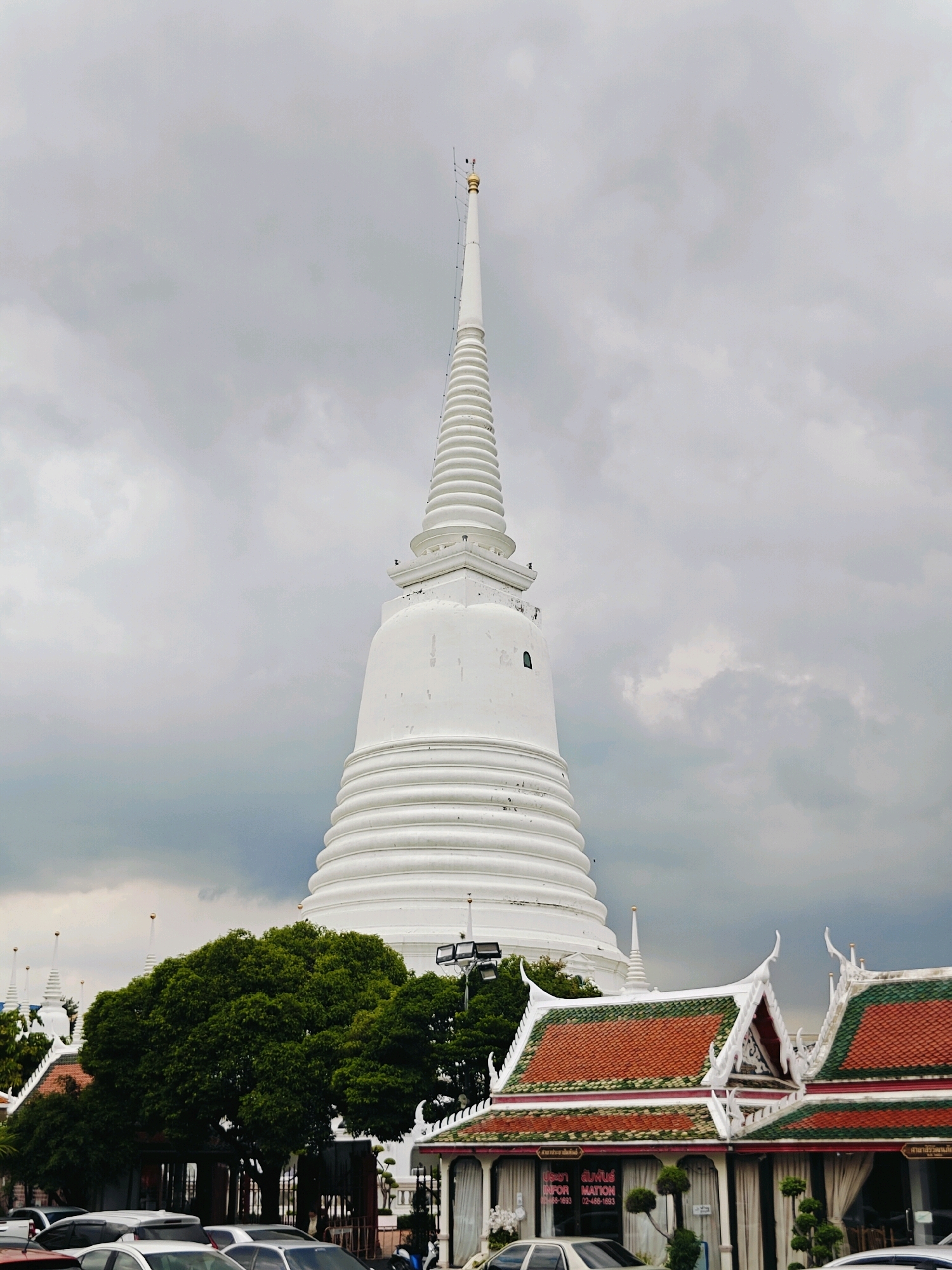 The image shows a view of Wat Intharawihan in Bangkok, Thailand. A large, white, tiered pagoda dominates the center of the frame, reaching towards a cloudy sky. The pagoda has a pointed spire topped with gold. In the foreground, the ornate, traditional Thai temple roofs with their distinctive swooping gables and green and orange tiles are visible. A dense, green tree stands between the temple roofs and the pagoda. Several cars are parked in the lower part of the image, suggesting a public space. The overall atmosphere is somewhat overcast, with muted lighting. There are no clearly discernible people in the image.