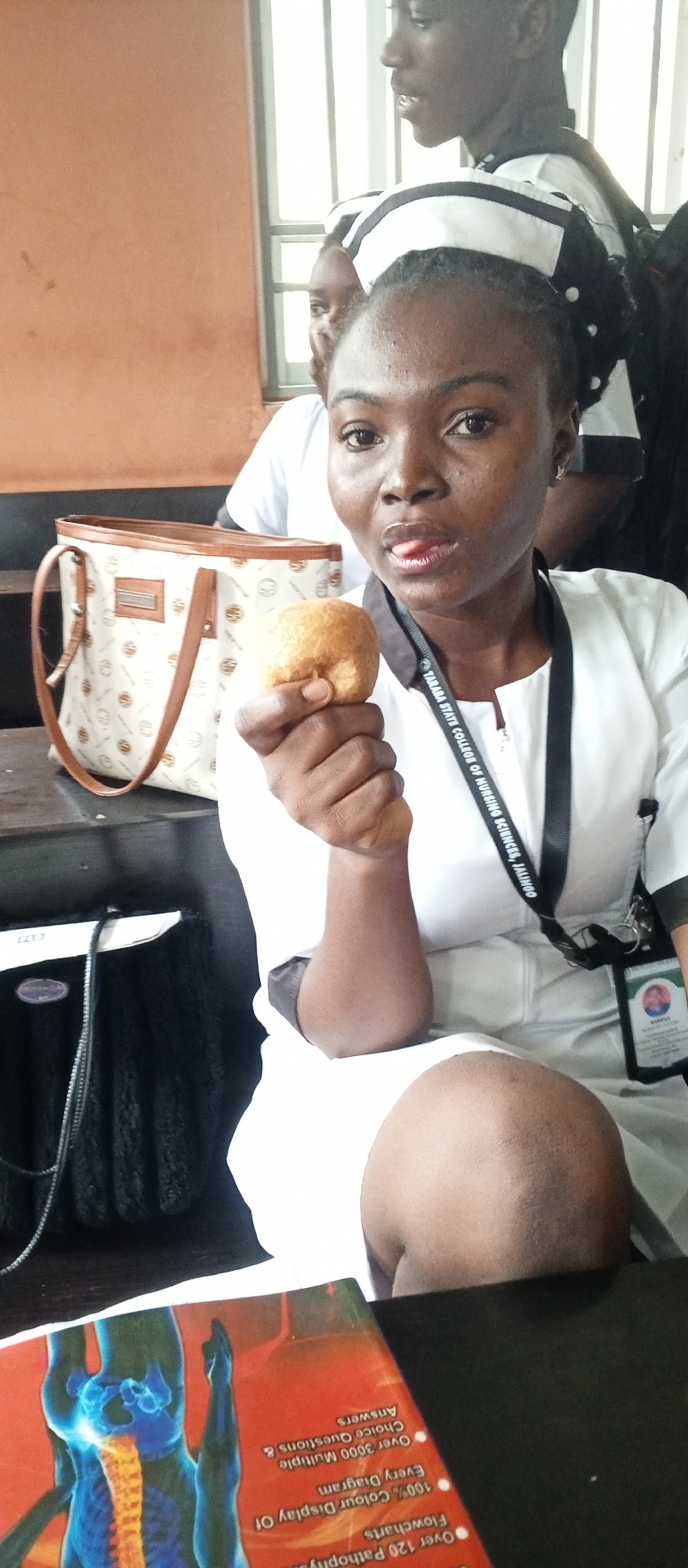 In Kuson, Nigeria, a young woman in a nurse's uniform, identified by a lanyard from Taraba State College of Nursing Sciences, Jalingo, is seated, holding a round, golden-brown pastry to her mouth with her right hand. Her expression is playful, with her tongue slightly extended. To her left, a light-colored handbag with a patterned design and brown trim rests on a dark surface. The background features a muted orange wall and a window with vertical bars. Other individuals in similar uniforms are partially visible behind her. In the foreground, a book with an anatomical illustration of the human spine is visible.
