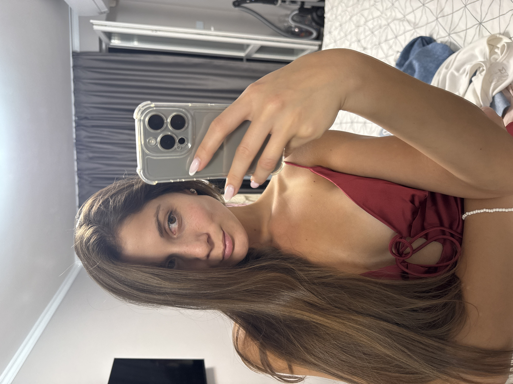 The image shows a young woman taking a mirror selfie in what appears to be a bedroom. She has long, brown hair and is wearing a red satin camisole. In her right hand, she holds a smartphone, its camera facing the mirror. The background reveals a bed with white and grey patterned bedding, dark grey curtains, and a window. To the left, a television screen is visible on the wall, and to the right, a vacuum cleaner stands near the window. The woman's posture and the setting suggest a personal, candid moment. The location is identified as Burgas, Bulgaria.