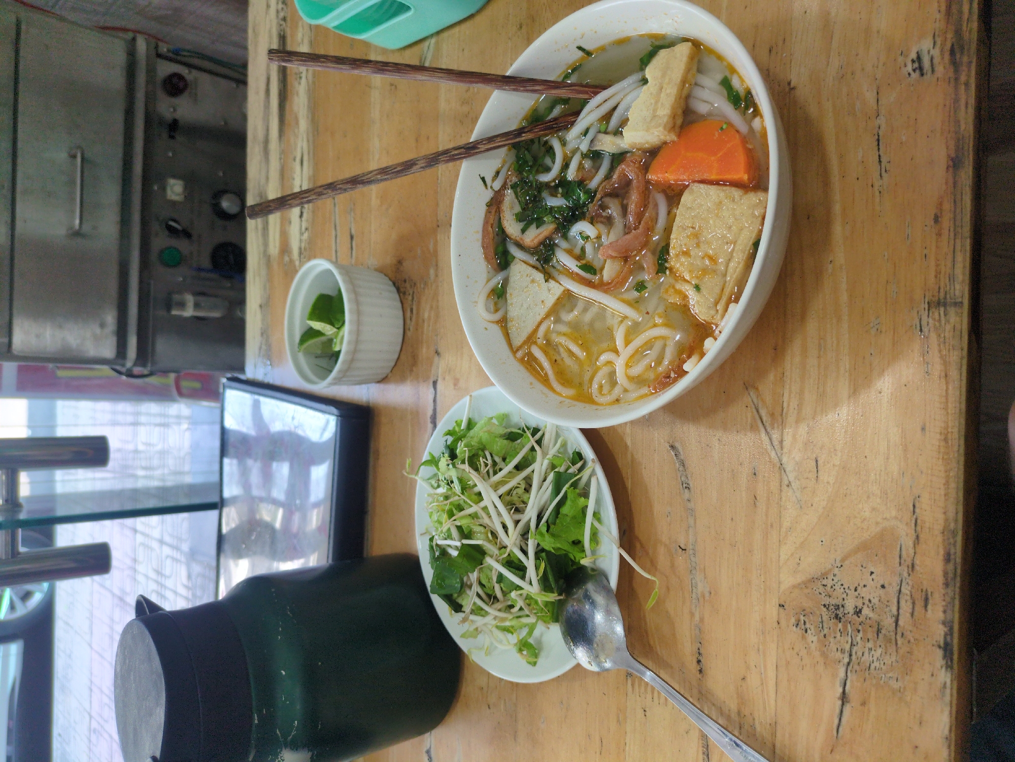 This is an eye-level, close-up shot taken in Nha Trang, Vietnam, showcasing a table laden with Vietnamese cuisine. The main focus is a bowl of what appears to be a Vietnamese noodle soup, possibly "Bun Cha Ca," characterized by thick white noodles, fried fish cake, a slice of carrot, and greens in a broth. Two chopsticks rest across the bowl. Beside the soup, there is a small white container holding lime wedges. A plate filled with fresh greens and bean sprouts lies near the soup, accompanied by a spoon. In the background, a section of a commercial kitchen appliance can be seen, hinting at the restaurant environment. The items are arranged on a light-brown wooden table. Adjacent to the food, a green water container and a small electronic device rest on a countertop near a glass display.