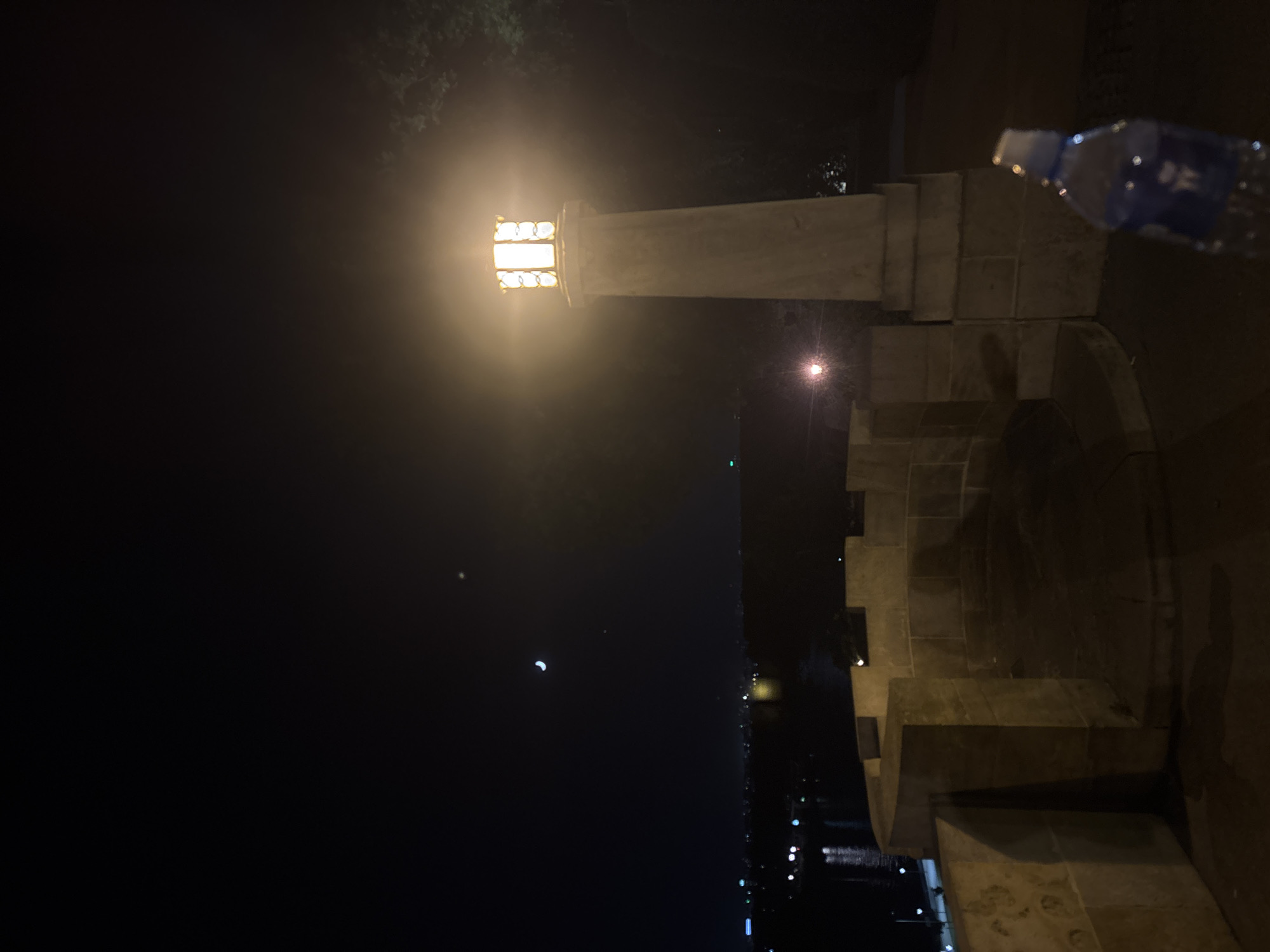Captured at night in Belgrade, Serbia, the image depicts a stone structure with a lit, ornate lamppost atop it. A bottle of water is visible to the right of the lamppost. In the background, faint lights suggest a cityscape along a body of water, likely the Sava or Danube River. A crescent moon can be seen in the dark sky.