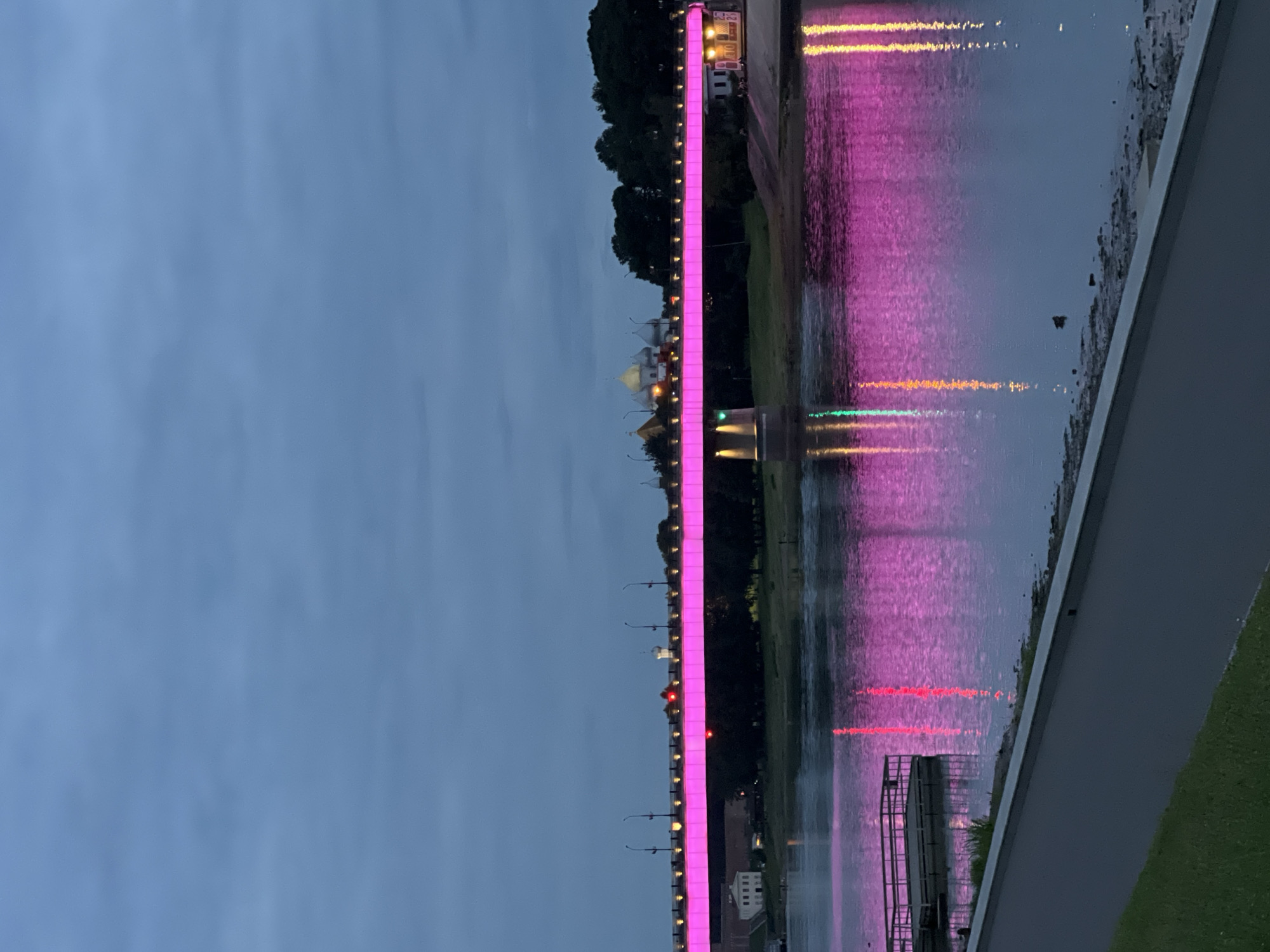 The image depicts a twilight scene of a bridge illuminated with pink lights over a body of water, likely the Volkhov River in Veliky Novgorod, Russia. The bridge stretches horizontally across the frame, with its pink lights casting reflections on the water's surface. On the far bank, trees and several buildings are visible, including what appears to be a church with a golden dome. A small dock or pier juts out into the water on the left side of the image. The sky is overcast and a deep blue, typical of dusk. The foreground shows a paved pathway adjacent to a grassy area.