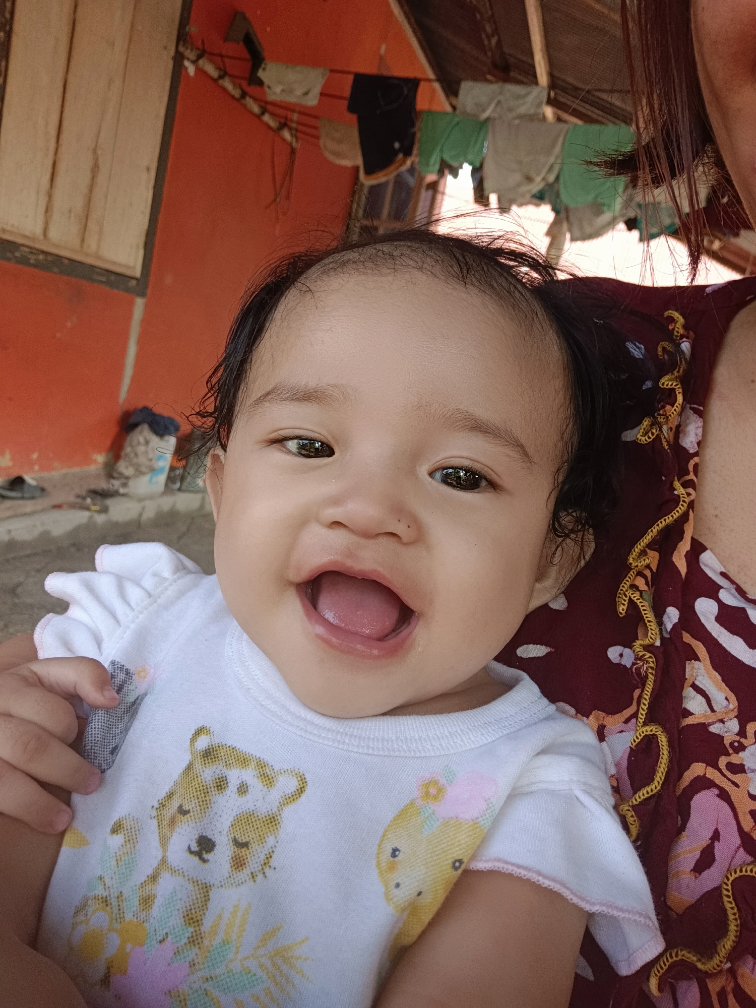 A close-up, eye-level outdoor portrait shows a smiling baby cradled by an adult, likely their parent. The baby, with dark hair and wide, dark eyes, is wearing a white t-shirt with a cartoon animal print. The adult, whose shoulder and upper chest are visible, is wearing a dark, patterned garment. The background is an exterior wall of a building with an orange hue, and to the left, a wooden structure is visible. Clothes hang on a line in the background, suggesting a laundry area. The ground appears to be concrete or dirt, with some debris visible. The overall scene suggests a casual, domestic setting, possibly in a village or rural area.