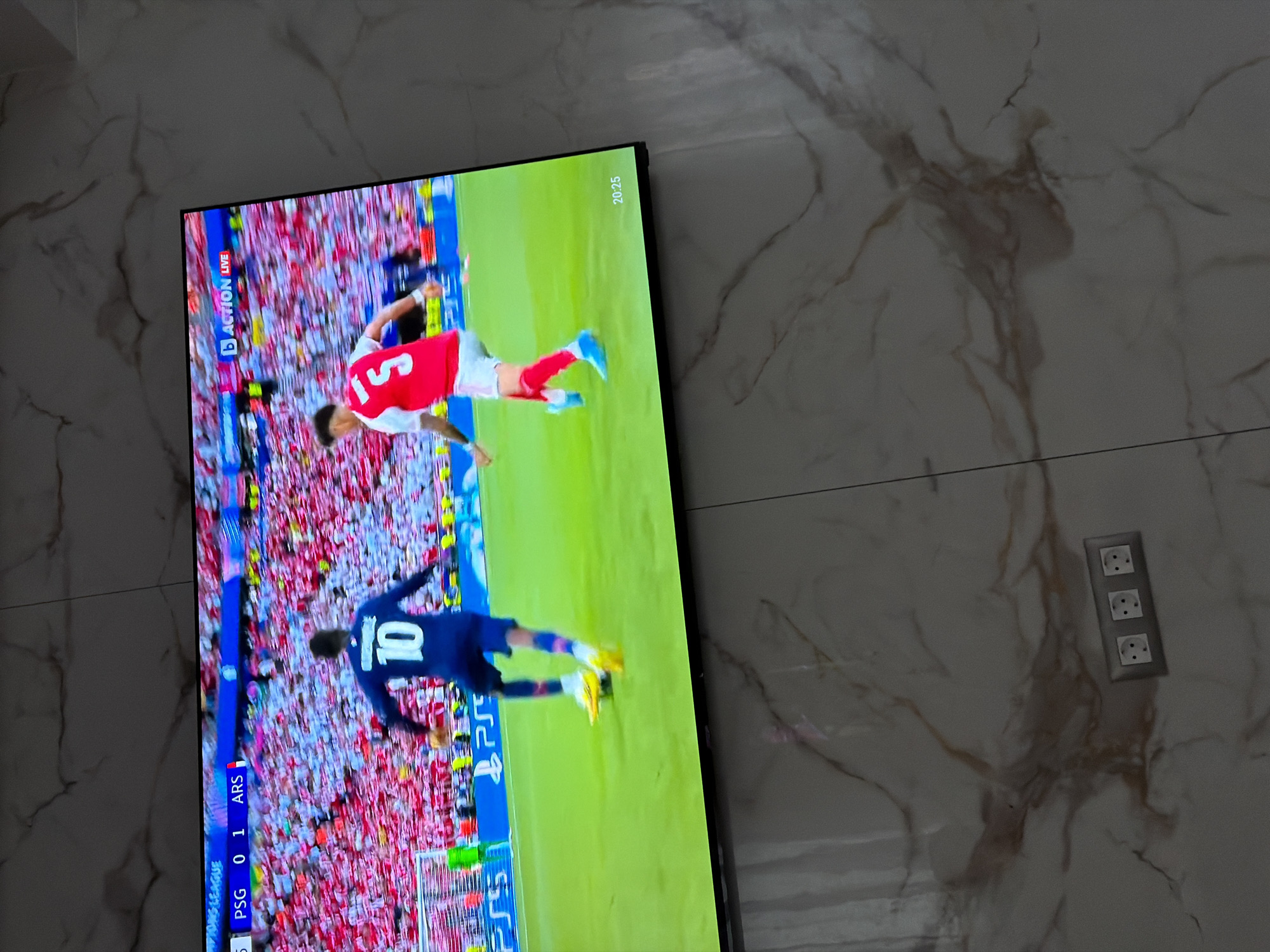 A television displaying a soccer game hangs on a marble wall. The score displayed on the screen shows PSG 0-1 ARS. In the top right corner of the screen, "20:25" is visible. The wall has a triple electrical outlet near the television. The location is likely a residence or business establishment in Asenovgrad, Bulgaria based on the type of electrical outlet.
