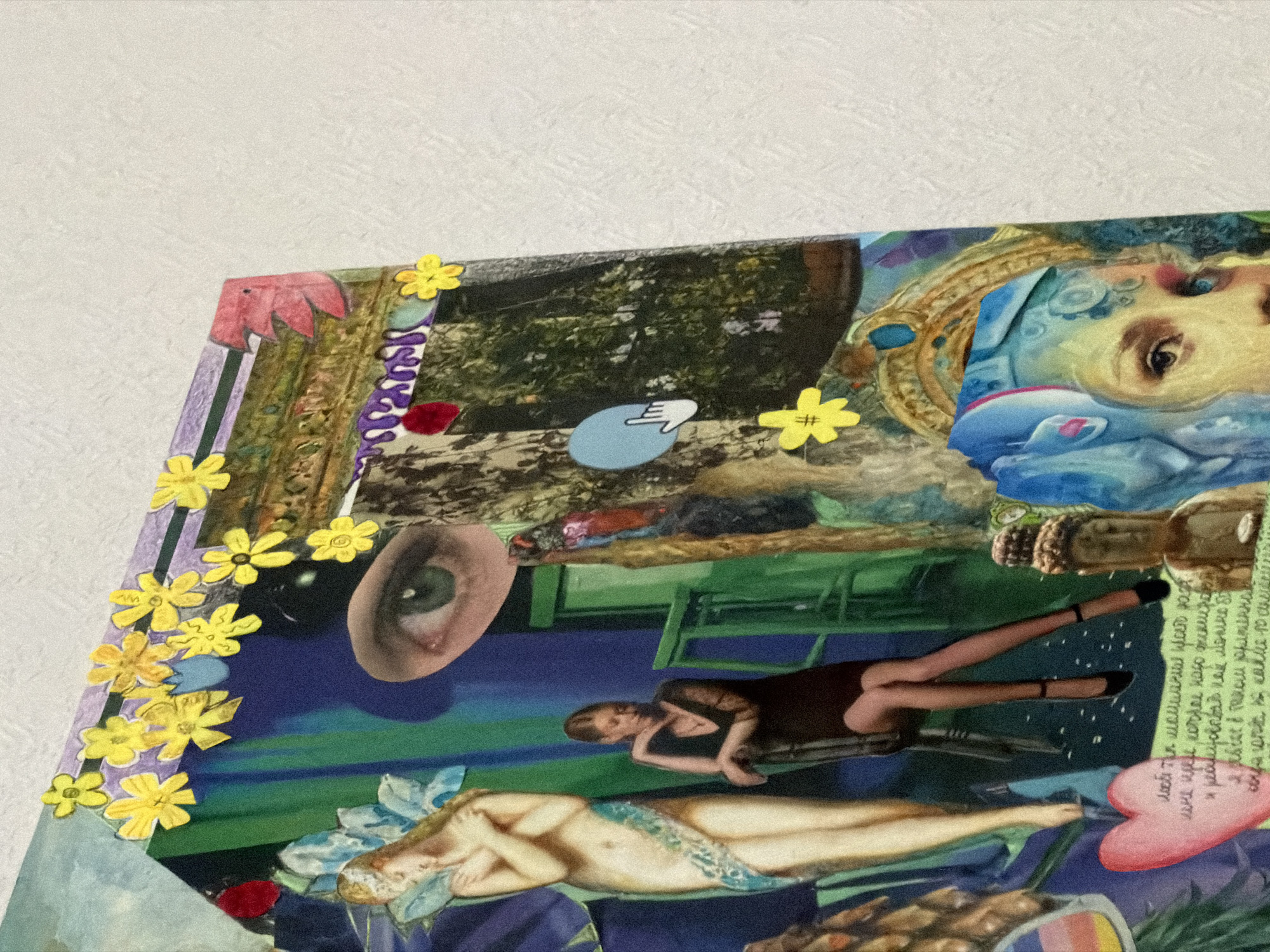 This is a close-up, angled, low-light photograph of a collage made of various cutouts and pasted materials, likely displayed on a flat surface. The collage features a mix of imagery including a classical statue of a nude female figure with blue accents, a woman in a black dress sitting on a chair, a Buddha figurine, a human eye, and numerous yellow paper flowers. Some of the collage elements appear to be hand-drawn or painted. There is text in an unintelligible script on a pink heart shape. The overall aesthetic suggests a surreal or dreamlike theme. The background of the image is a textured, off-white surface, possibly a wall. There is no identifiable information within the image to suggest a specific location in Moscow, Russia.