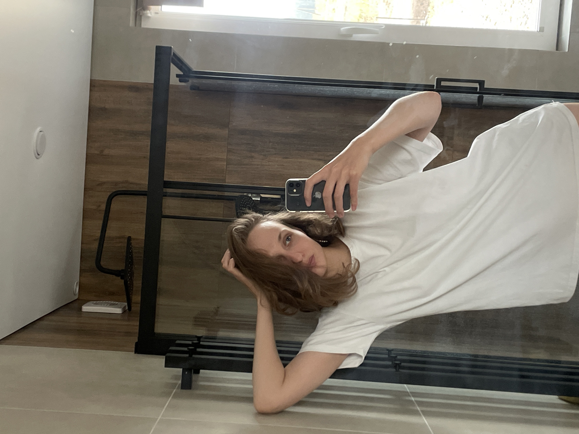 The image appears to be a selfie taken in a bathroom, possibly in Подшивалово, Russia. A young woman with shoulder-length brown hair is holding a smartphone and taking the picture in a mirror. She is wearing a white t-shirt. The bathroom has light-colored tile flooring, and the walls have a mix of light-colored paint and brown wood paneling. A black metal towel rack or shelf unit is visible in the reflection. There is also a window with natural light coming through. A white object, potentially a scale or bathroom accessory, is visible on the floor near the wall.