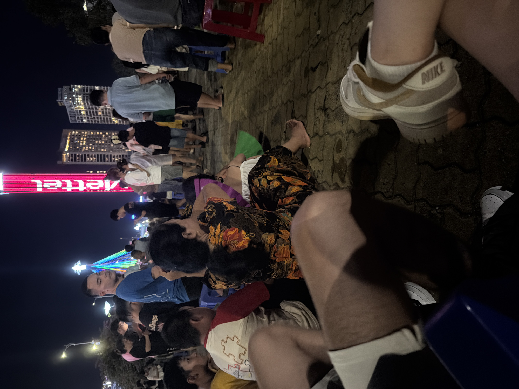 The image depicts a crowded nighttime scene in Đà Nẵng, Vietnam, likely along a public promenade or event space near the Han River.
In the background, the dark night sky is illuminated by artificial light sources. A prominent, tall, red-illuminated tower, displaying the vertical "Viettel" logo, stands on the right. To its left, several modern high-rise buildings with numerous illuminated windows contribute to the urban skyline. A multi-colored, conical or pyramidal light structure, possibly an artistic installation or festive decoration, glows vibrantly to the left of the high-rise buildings. Standard streetlights provide additional illumination.
The mid-ground features a dense gathering of people, mostly adults, engaged in various activities. Some individuals are standing and observing the surroundings, while others are seated on small plastic stools or directly on the paved ground. A man in a blue t-shirt with short dark hair stands prominently in the center-left, looking directly towards the camera. To his right, another man in a light blue polo shirt and dark shorts walks away from the viewer. Further right, a man in a tan t-shirt and blue jeans stands with his back partially to the camera. In the center-right mid-ground, an individual with dark hair tied in a ponytail is seated on the ground, facing away from the camera, wearing a dark shirt patterned with orange, red, and yellow flowers, and patterned dark pants. Next to them, another person is seated, wearing a white raglan shirt with red sleeves and a large, multi-colored puzzle piece graphic on the back. Small blue and red plastic stools are visible as seating for some attendees.
In the immediate foreground, the lower bodies of two individuals are visible. On the left, a person's bare legs are seen, wearing white shorts and dark pants, with white sneakers partially visible. On the right, another person's bare legs are shown, wearing white socks and a light-colored Nike sneaker with tan accents.
The ground is paved with interlocking concrete blocks, appearing somewhat damp or reflective under the ambient light. The overall scene suggests a public event or festive gathering in a bustling urban environment.
