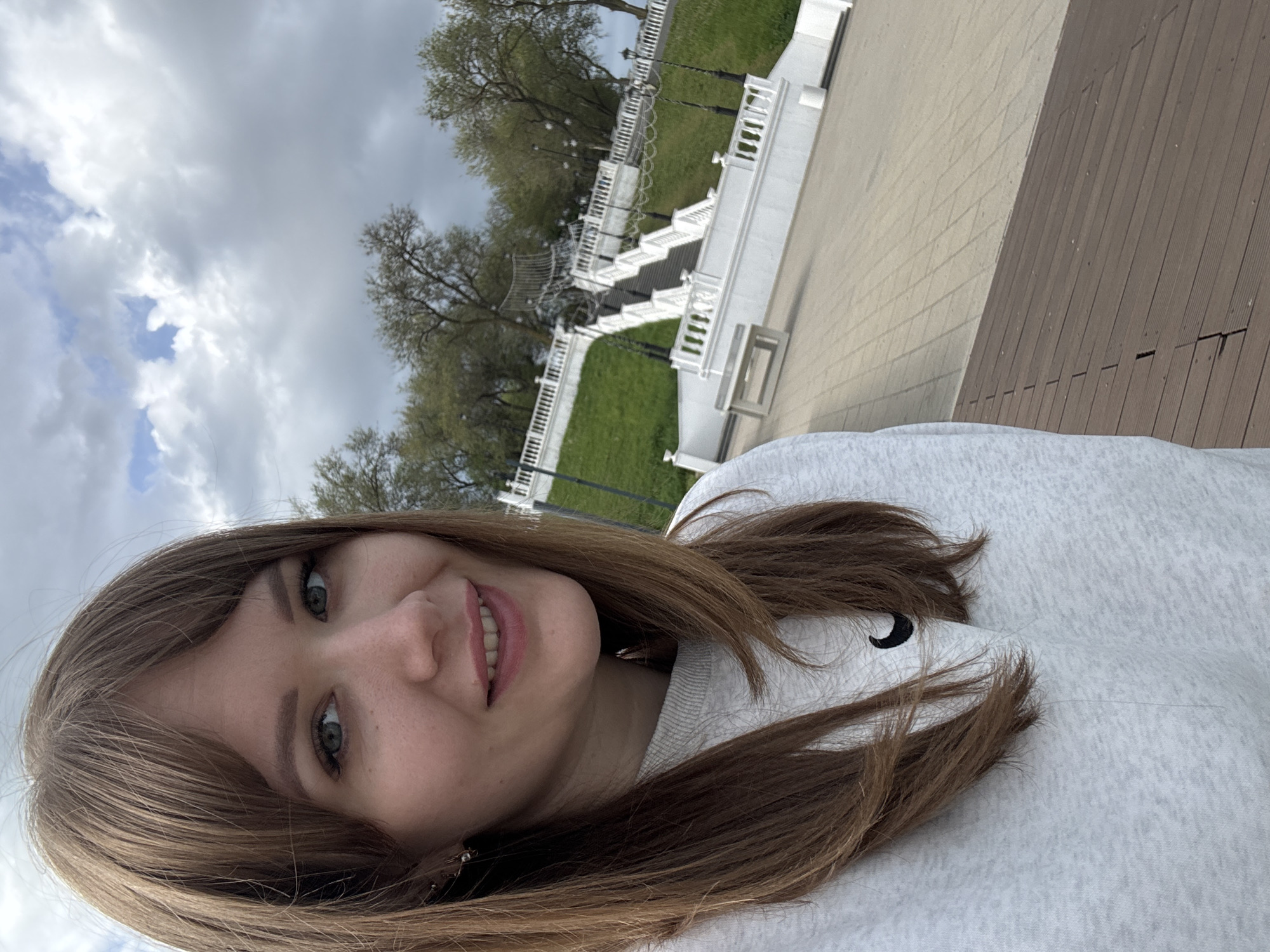 A selfie was taken outdoors, featuring a young woman with shoulder-length brown hair and green eyes. She is wearing a light gray sweatshirt. The background reveals a cloudy sky, trees, a white decorative staircase, a green grassy area, and a wooden platform. The location is Taman, Russia.