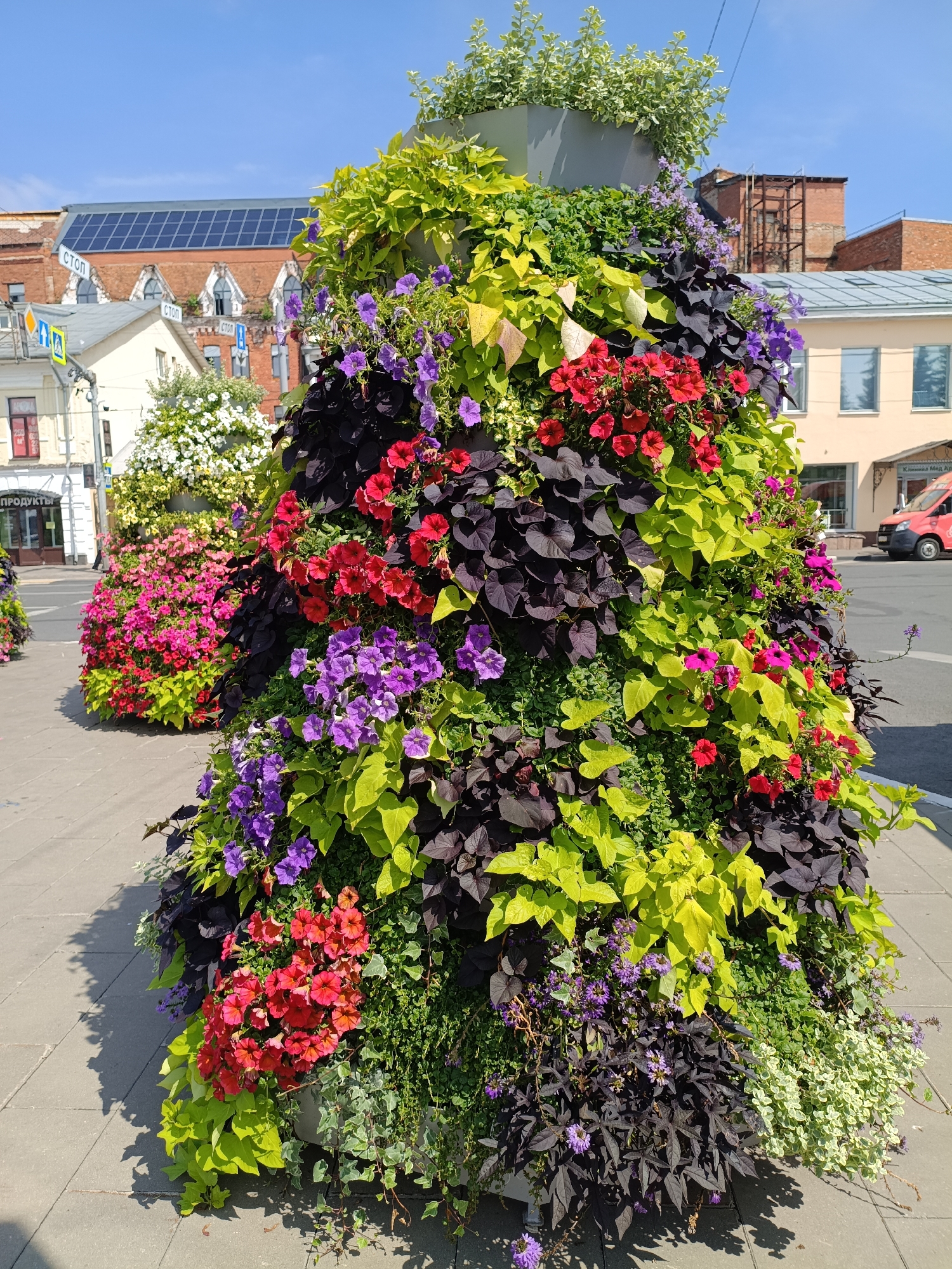 This image captures a vibrant street scene in Yaroslavl, Russia, on a sunny day. The foreground is dominated by an elaborately decorated, conical floral display that cascades with a variety of colorful flowers and lush green foliage. Red, purple, and pink petunias are interspersed with lime green and deep purple trailing vines, creating a rich tapestry of color and texture.
Behind the floral installation, a paved sidewalk leads to a street. In the middle ground, another similar, though less detailed, floral arrangement can be seen. Further back, buildings line the street, showcasing architectural styles typical of Russian cities. One building, to the left, has a sign with Cyrillic lettering that appears to indicate "PRODUKTY," suggesting a shop. Another building to the right features solar panels on its roof. A red vehicle is visible on the street. The sky above is a clear, bright blue. The overall impression is one of urban beautification, with the floral arrangements adding a significant splash of nature to the cityscape.