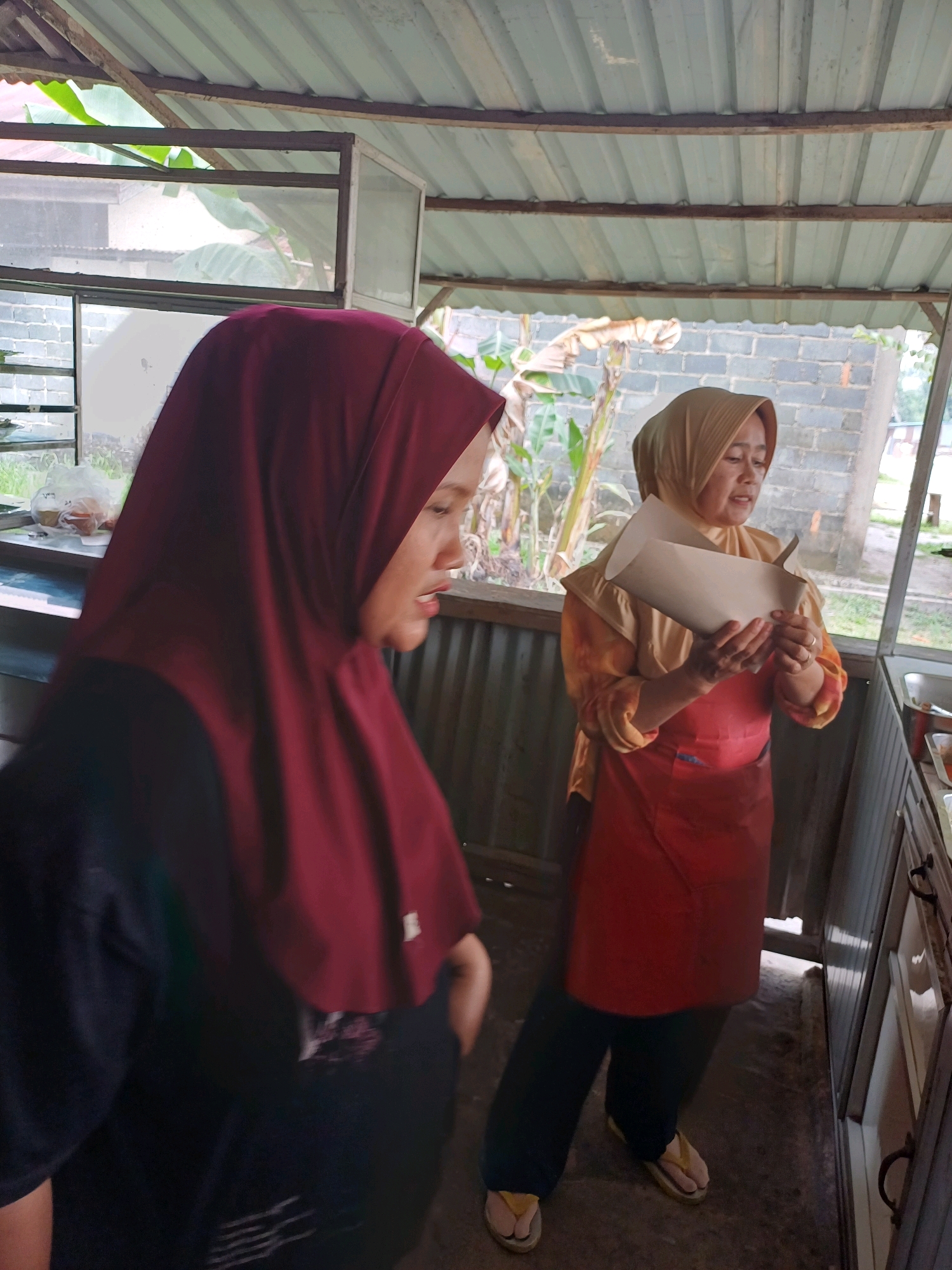 Two women are inside a food stall in Dumai City, Indonesia. The woman on the left is wearing a dark blue t-shirt and a maroon hijab. She is partially visible and facing away from the camera. The woman on the right is wearing a yellow patterned shirt, a red apron, and a light brown hijab. She is holding a paper container and appears to be speaking. The food stall has a corrugated metal roof and is open to the outside. Various food preparation items and ingredients are visible in the background. The ground is concrete.