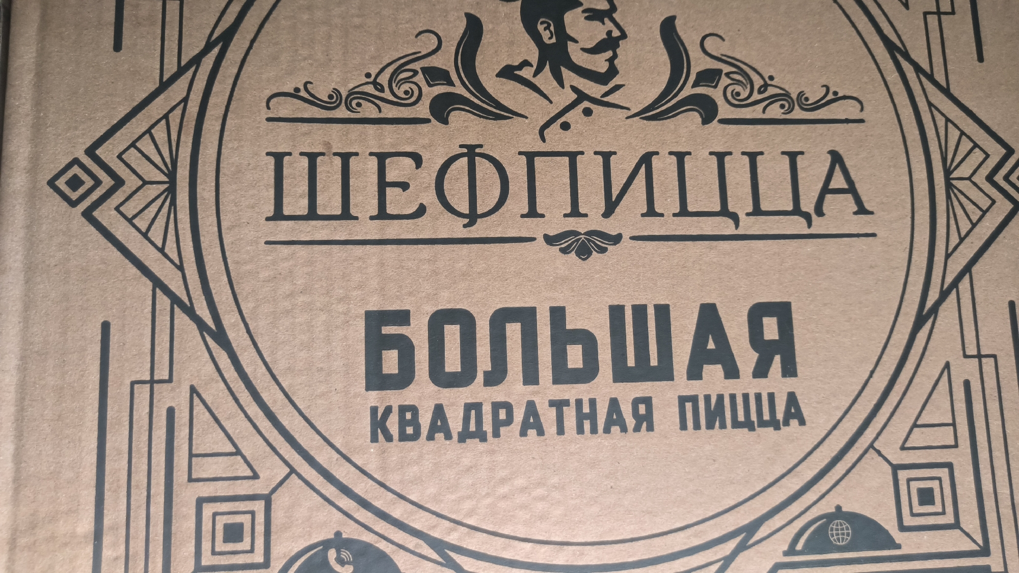The image shows a close-up of a cardboard box with a stylized, dark brown design and Cyrillic text. The design features a circular motif with art deco-style elements on the sides, including geometric shapes and lines. At the top center of the circle is an illustration of a man's head in profile with a mustache and hat, reminiscent of a chef.
The text within the circle reads:
"ШЕФПИЦЦА" (SHEFPIZZA) in a large, serif font.
"БОЛЬШАЯ" (BOLSHAYA - meaning "large") in a very large, bold, sans-serif font.
"КВАДРАТНАЯ ПИЦЦА" (KVADRATNAYA PIZZA - meaning "square pizza") in a smaller, sans-serif font below "БОЛЬШАЯ".
There are no people or discernible actions in the image, only the packaging of a product. The background is the textured surface of the cardboard. The image is likely a product shot of pizza packaging. There is no information within the image that would allow for its specific location in Kovrov, Russia.