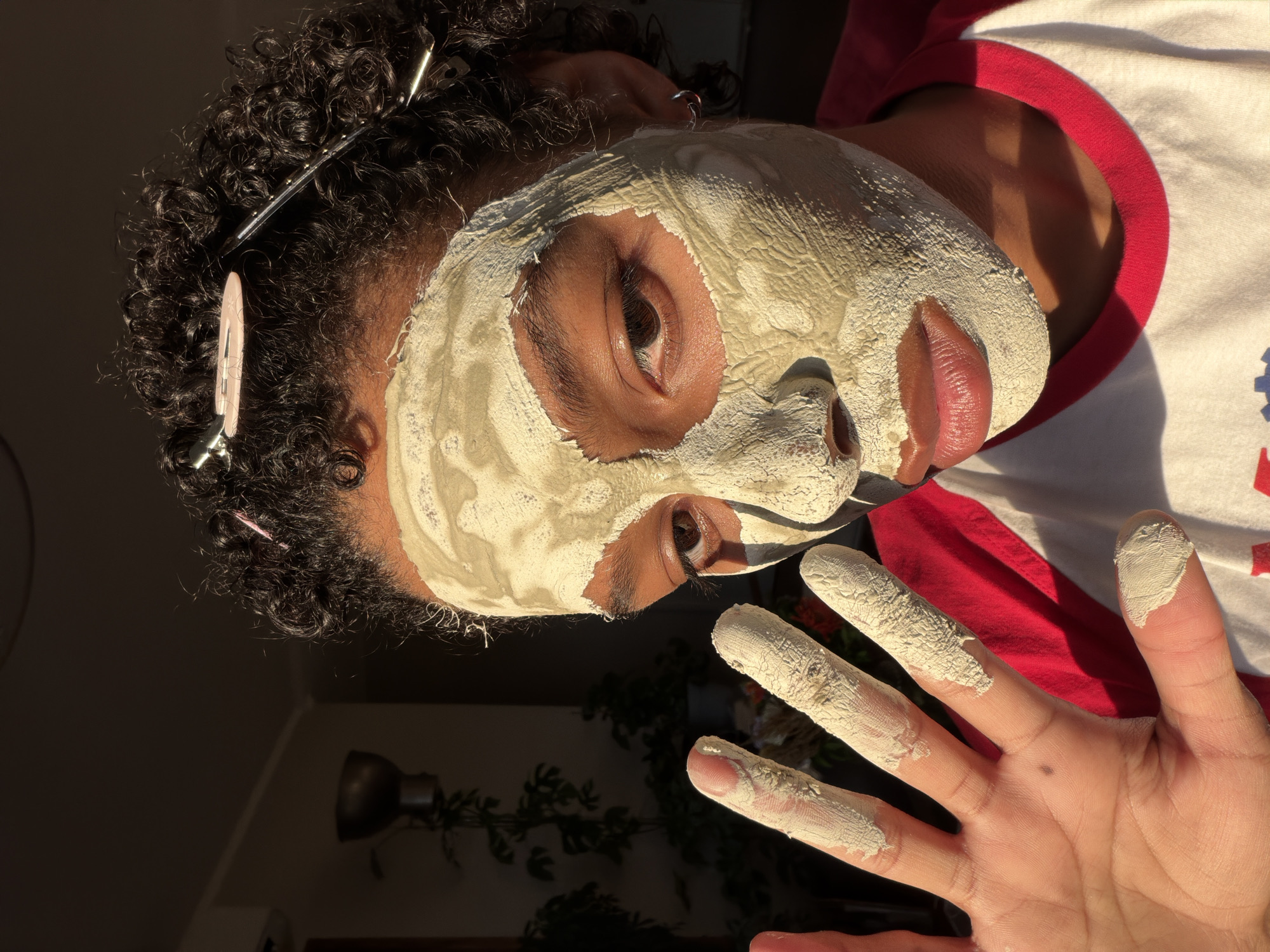 A person with curly dark hair, secured with two hair clips, applies a light green clay mask to their face. The mask covers the majority of their face, leaving only the eyes and lips visible. Their right hand, covered in the same clay mask, is held up towards the camera, with fingers splayed. The person is wearing a white t-shirt with a red collar. The background is dimly lit, with a plant visible to the left and a lamp on the wall. The scene is illuminated by warm, directional light, casting shadows. The person's skin appears to be of a medium complexion. The location is the city of Champs-sur-Marne, France.