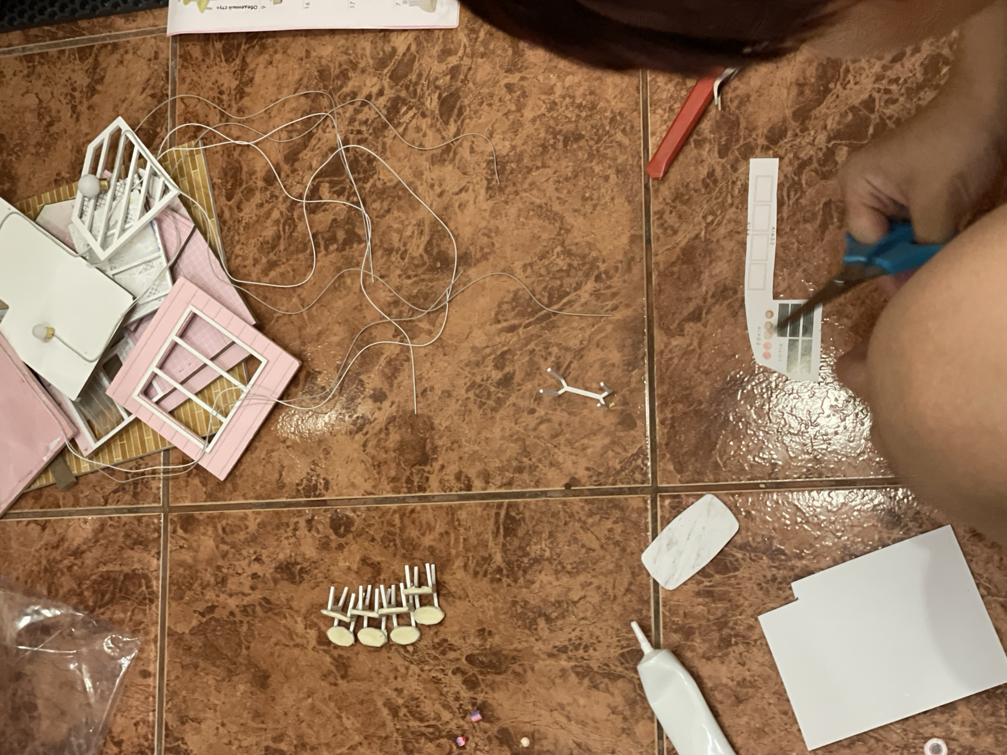 The image displays a close-up, high-angle view of a person's hands assembling a miniature dollhouse kit on a tiled floor. The floor is a brown, marble-patterned tile. Scattered on the floor are various components of the dollhouse kit, including pink and white plastic pieces, a white tube of glue, small beads, a piece of paper with small printed squares, and a pair of blue scissors held by the person. A red cutting tool is also visible. The person's legs and lower torso are partially in view. The lighting suggests an indoor setting, with the light source appearing to be from above and slightly to the right. There is no information in the image to suggest the location is in Sochi, Russia.