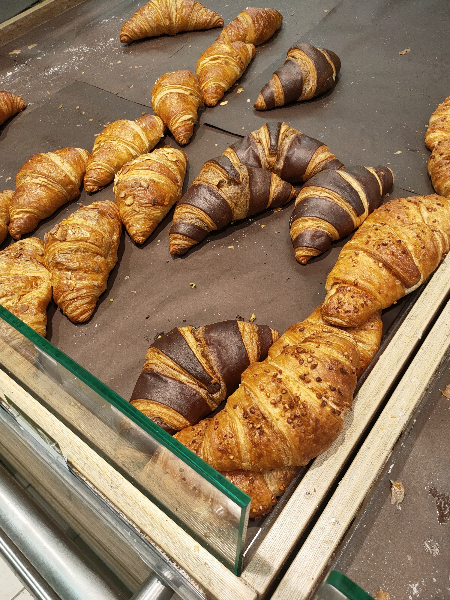 The image displays a variety of freshly baked croissants arranged in a display case. The croissants are of different varieties, including plain, chocolate, and almond-topped. The display case has wooden dividers and a glass front, suggesting it is in a bakery or a food establishment. The lighting appears to be artificial, typical of an indoor setting. The scene is static, with no people or specific activity visible, focusing solely on the food items. The overall impression is one of abundance and temptation, showcasing a selection of pastries ready for purchase. The context of Kyiv, Ukraine, is provided as a location cue, but there are no overt visual indicators in the image itself to confirm this specific location.