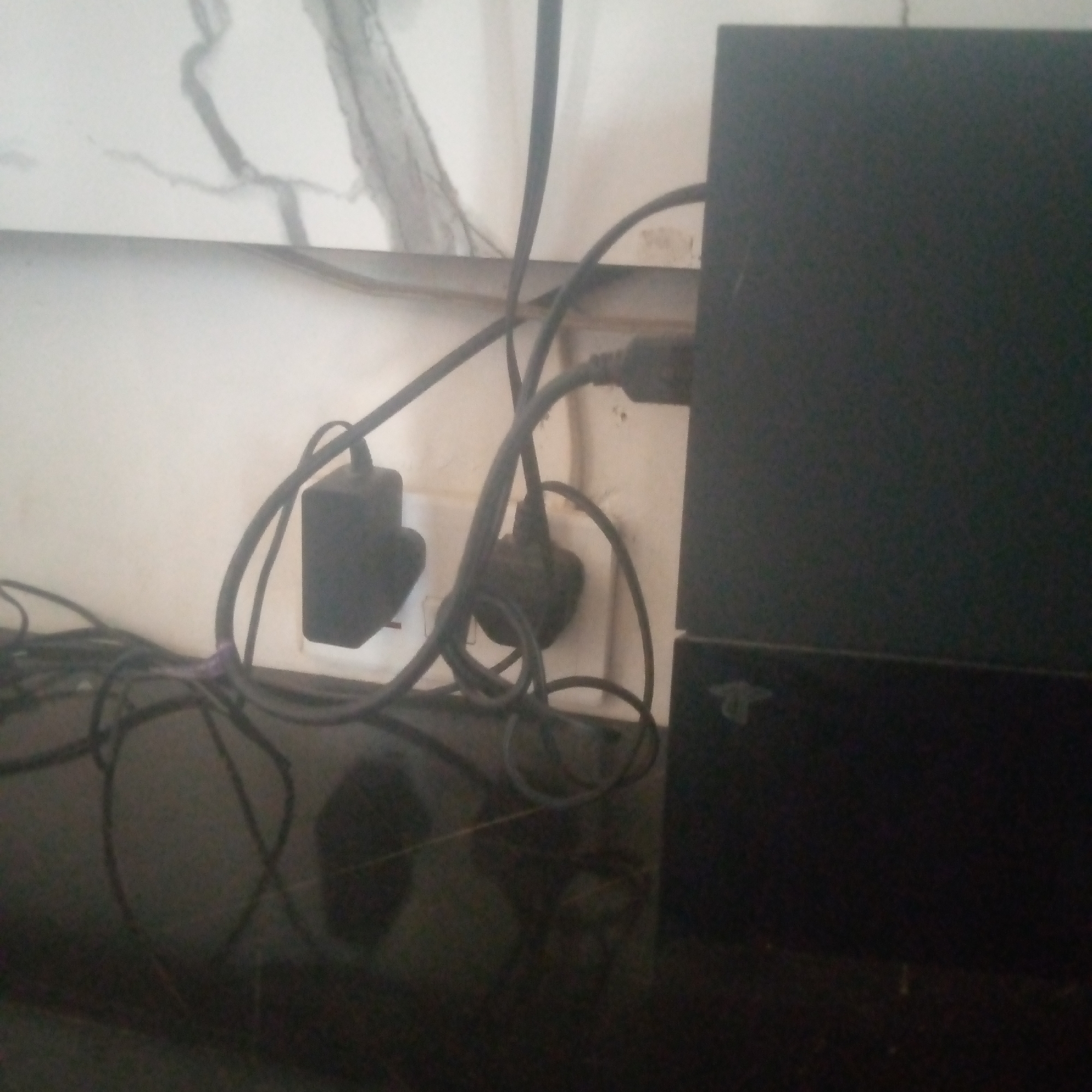 The scene, located in the city of Unknown, Unknown, presents a static, medium-shot view of an electronic setup in an indoor environment. A black Sony PlayStation console, identified by its distinct form factor and visible logo on the lower unit, is positioned on the right side of the frame, resting on a dark, reflective planar surface. Behind the console, a dense network of black electrical cables is visible, with one section bundled by a purple tie. These cables connect to two wall-mounted electrical outlets on a light-colored wall, which exhibits faint grey vein-like patterns. A black rectangular power adapter is plugged into the leftmost outlet, and another black power plug is inserted into the outlet positioned to its right. No individuals are present in the image. The overall lighting is dim, and the image displays a slight blur.
