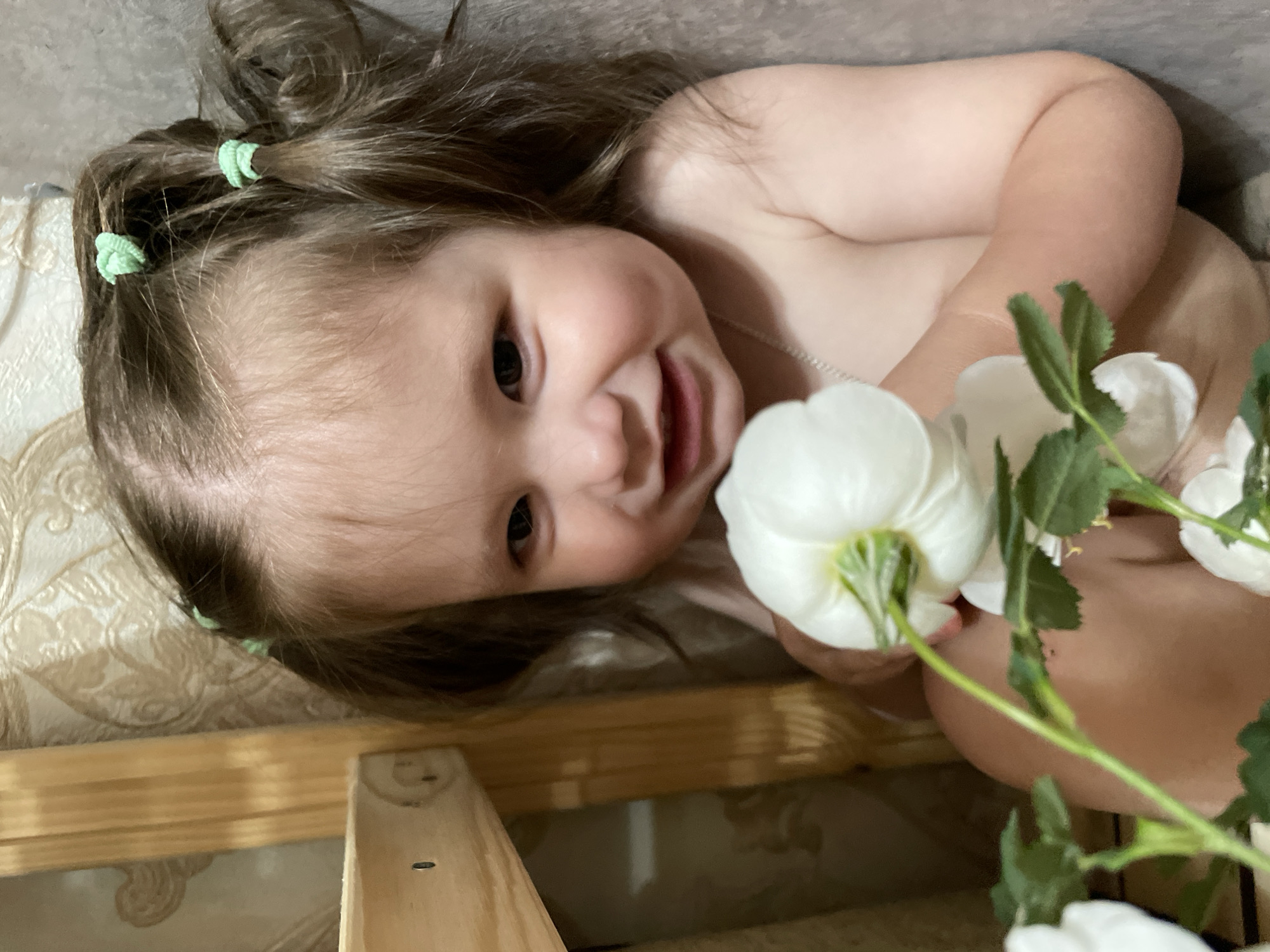 A close-up, low-angle shot depicts a baby lying on a beige surface, likely indoors. The baby has fair skin, brown hair, and green hair ties holding back sections of their hair. They are smiling and holding white flowers with green stems and leaves. The location cannot be determined from the image; it is not possible to ascertain if this image was taken in Izhevsk, Russia. The surrounding environment appears to be a wooden structure with a patterned surface beneath.
