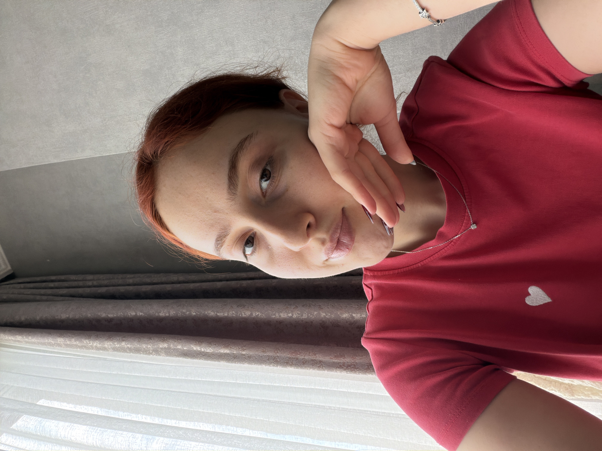 The image is a sideways-oriented, close-up selfie of a young woman taken indoors, likely during the daytime in Tbilisi, Georgia. The subject, a woman with light skin and reddish-brown hair, occupies the majority of the frame. She looks directly at the camera with a calm and gentle expression, possibly a faint smile, revealing subtle freckles across her face. Her left hand is raised to her cheek and temple, with fingers slightly spread. Her fingernails are painted a dark, possibly purplish-grey shade.
She is wearing a simple, short-sleeved red t-shirt, which features a small, light-colored heart emblem embroidered on the left chest area. A delicate silver chain necklace with a small pendant is visible around her neck, resting on her collarbone. A silver bracelet with intricate details can be seen on her right wrist, partially obscured by her arm.
The background consists of interior elements. To the left, a light grey ceiling or upper wall is visible. Below that, a combination of window coverings can be seen: darker, possibly textured curtains or blinds at the top, layered over lighter, sheer curtains or blinds that allow soft natural light to pass through. The overall lighting suggests it is daytime. There is no visible text in the image.