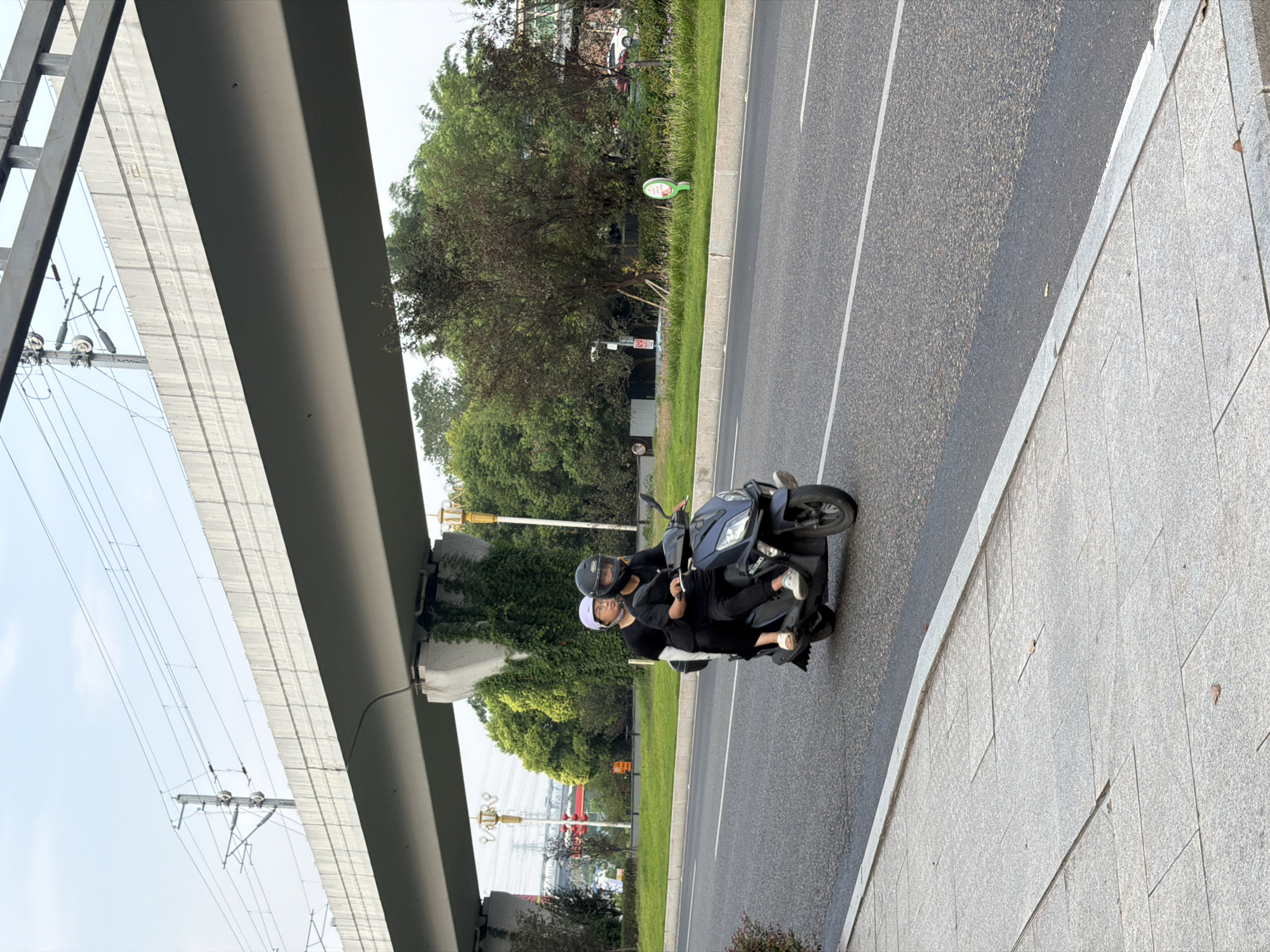 Two individuals are riding a dark blue electric scooter on a paved road. The person in front, wearing a black t-shirt and helmet, is operating the scooter. The person behind, wearing a black t-shirt and a light purple helmet, is a passenger. They are both wearing casual clothing and appear to be traveling on a multi-lane road with a concrete barrier on the left and a grassy area with trees and streetlights on the right. Above the road, a large elevated structure, likely a highway or railway bridge, spans across the scene, supported by concrete pillars covered in greenery. Power lines are visible above the elevated structure. The overall environment suggests an urban or suburban setting in the city of Jiangjin, China.