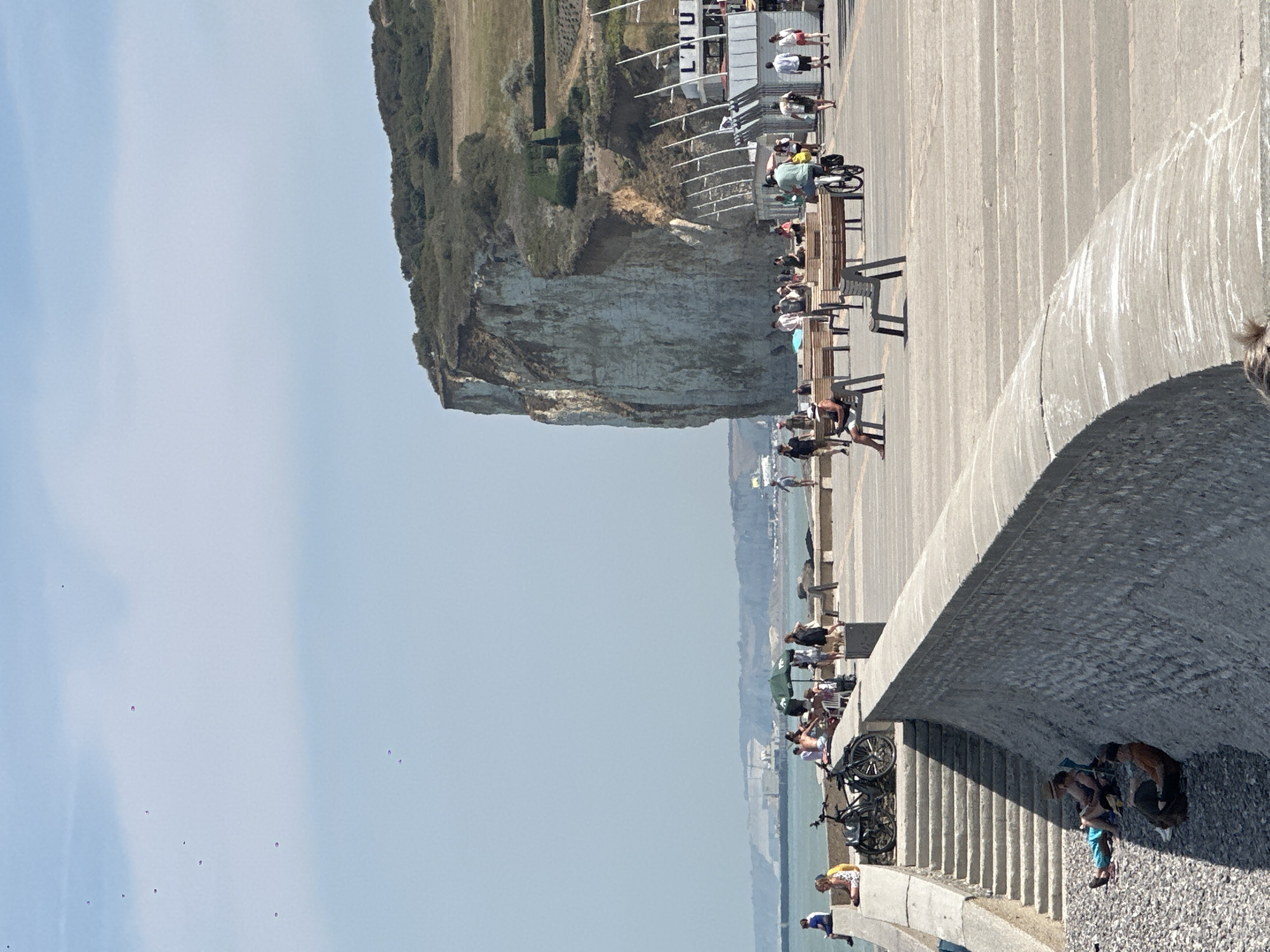 The image depicts a coastal scene in Hautot-sur-Mer, France, under a clear, pale blue sky. In the foreground, a stone wall curves, with a set of concrete stairs leading up from a pebble beach where three individuals are seen relaxing on the ground. Above the wall, a wide, paved promenade is populated with people. Some are sitting on benches, others are walking, and a person is riding a bicycle. Bicycles are also parked near the stairs.
Dominating the mid-ground and background is a large, steep, white chalk cliff, characteristic of the Alabaster Coast. The cliff face is textured and appears to be covered in vegetation at its upper reaches, with a sloping, grassy area above. Buildings with white facades and vertical supports are visible at the base of the cliff, suggesting a developed area or amenities. Further in the distance, a hazy coastline stretches out, with more cliffs and structures visible. The sea is a muted blue-grey and appears calm. Several small, dark objects are scattered in the sky, possibly birds or airborne debris. The overall impression is of a sunny, recreational day at the seaside.