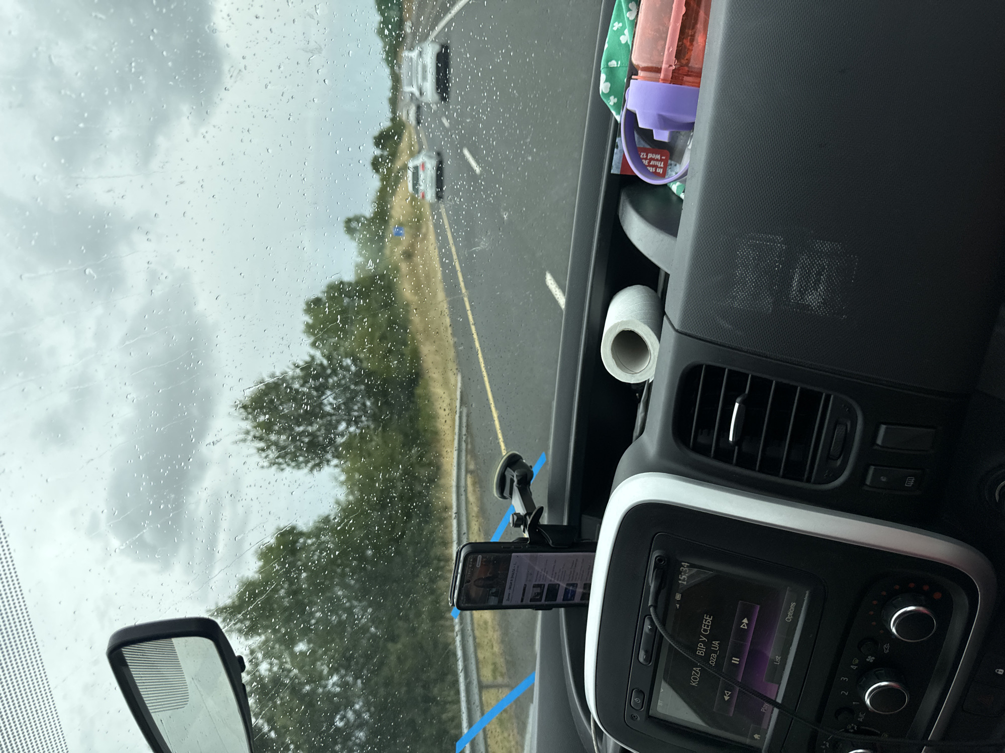 The image shows a view from inside a vehicle looking out through a rain-streaked windshield. The vehicle is on a highway, with other cars visible in the distance. The sky is overcast and it appears to be raining. The interior of the vehicle is visible in the foreground, including a dashboard with a car stereo, air vents, and a phone mounted on a stand. There are also some personal items on the dashboard, such as a roll of toilet paper and a water bottle. The image does not provide any information about the location in the city of Craan, Ireland, or any specific details about people, subjects, actions, or interactions.