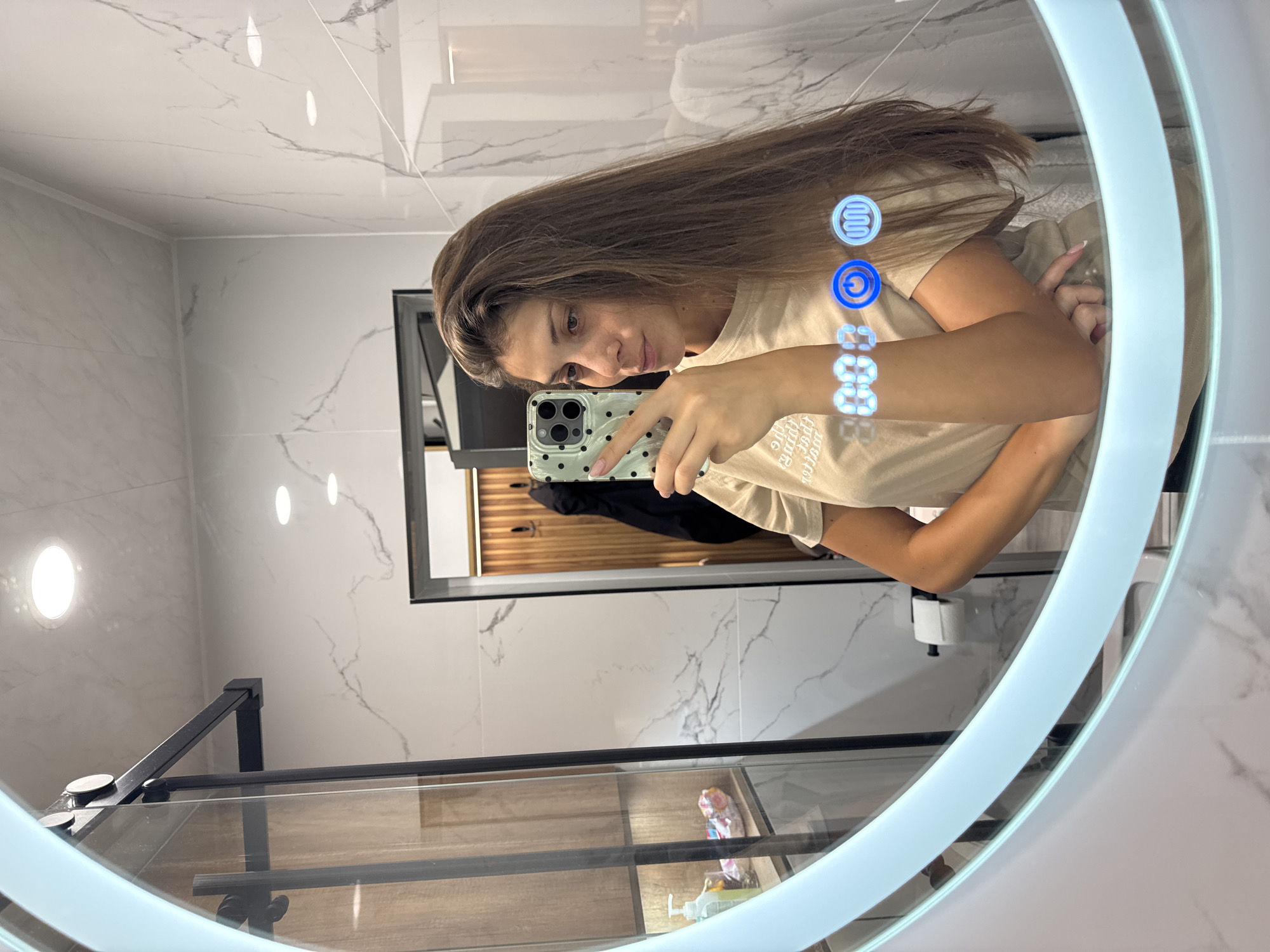 The image shows a woman taking a selfie in a bathroom mirror. The bathroom has white tile walls. The woman has long brown hair and is wearing a beige t-shirt. She is holding a smartphone with a light green case with black dots. In the background, there is a wood-paneled wall with a dark jacket hanging on it. The image is taken inside, likely a hotel or residence. There is no explicit evidence to confirm that the scene is in Burgas, Bulgaria.