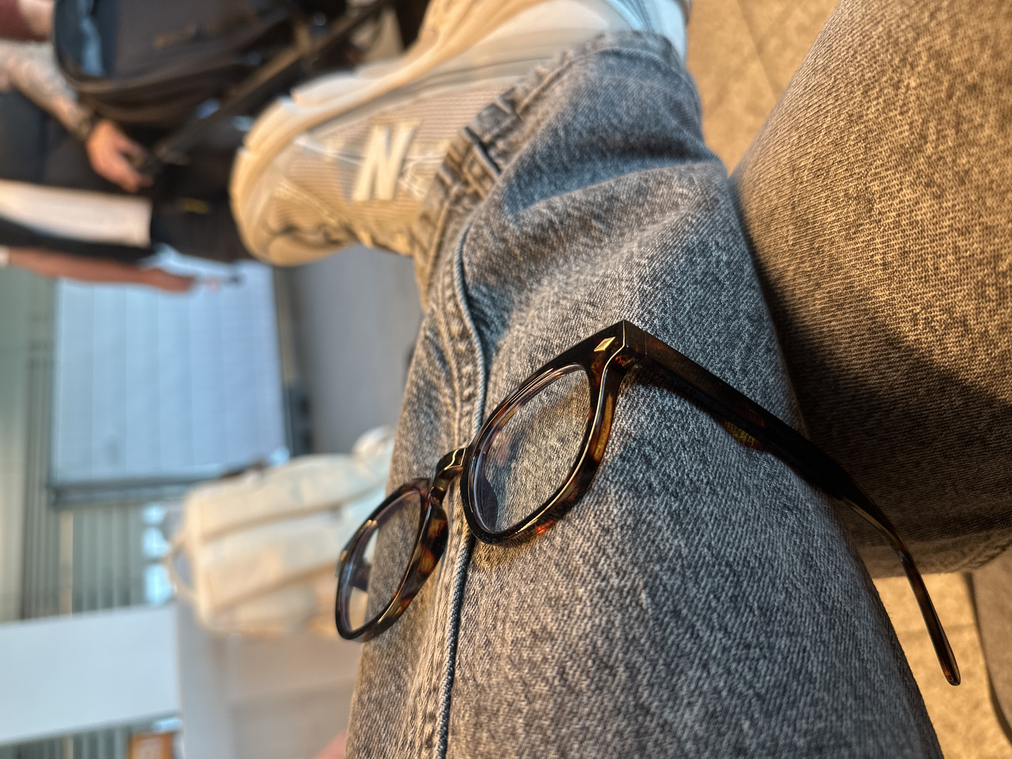 The image presents a close-up, angled perspective focusing on a pair of tortoiseshell eyeglasses resting on a person's leg, which is clad in textured grey denim jeans. The eyeglasses have a classic, rounded frame with a distinctive mottled brown and amber pattern.
In the upper left portion of the frame, the top of a light-colored sneaker is visible on the same leg, featuring a prominent white "N" logo, indicative of a New Balance shoe. The background is softly blurred but reveals elements typical of an indoor public waiting area. To the left, a shadowy figure, likely another person, is partially visible with what appears to be a dark backpack and an arm displaying a tattoo. To the right, a light-colored, possibly white, piece of luggage or a bag is indistinctly seen.
The setting appears to be a spacious indoor environment with bright, indirect lighting, suggesting a daytime scene, possibly within an airport or a large transport terminal, consistent with the location context of Weeze, Germany. The overall atmosphere is relaxed, with the subject seemingly resting or waiting. No specific activity beyond sitting and waiting is clearly depicted. No other explicit text is visible besides the "N" logo.