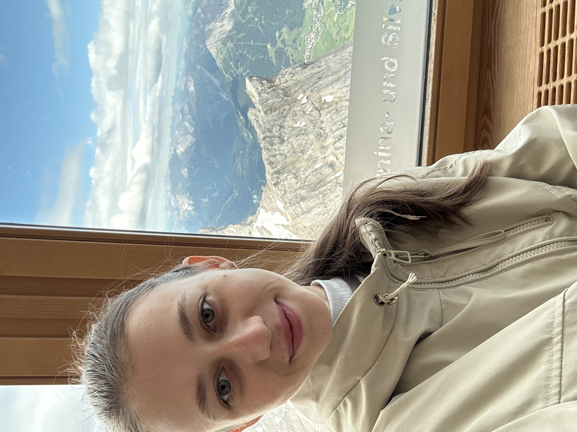 A young woman with brown hair tied in a ponytail takes a selfie in a room with a large window offering a panoramic view of the Zugspitze mountain range in Ehrwald, Austria. The woman is wearing a light beige hooded jacket over a light blue turtleneck sweater. Her expression is a slight smile. The window behind her shows a vast expanse of rugged, snow-capped mountains under a partially cloudy blue sky, with a small village visible in the valley below. A wooden frame surrounds the window, and a metal grate is visible on a wooden bench in the foreground. Text is visible on the window, reading "nina- und Silv".