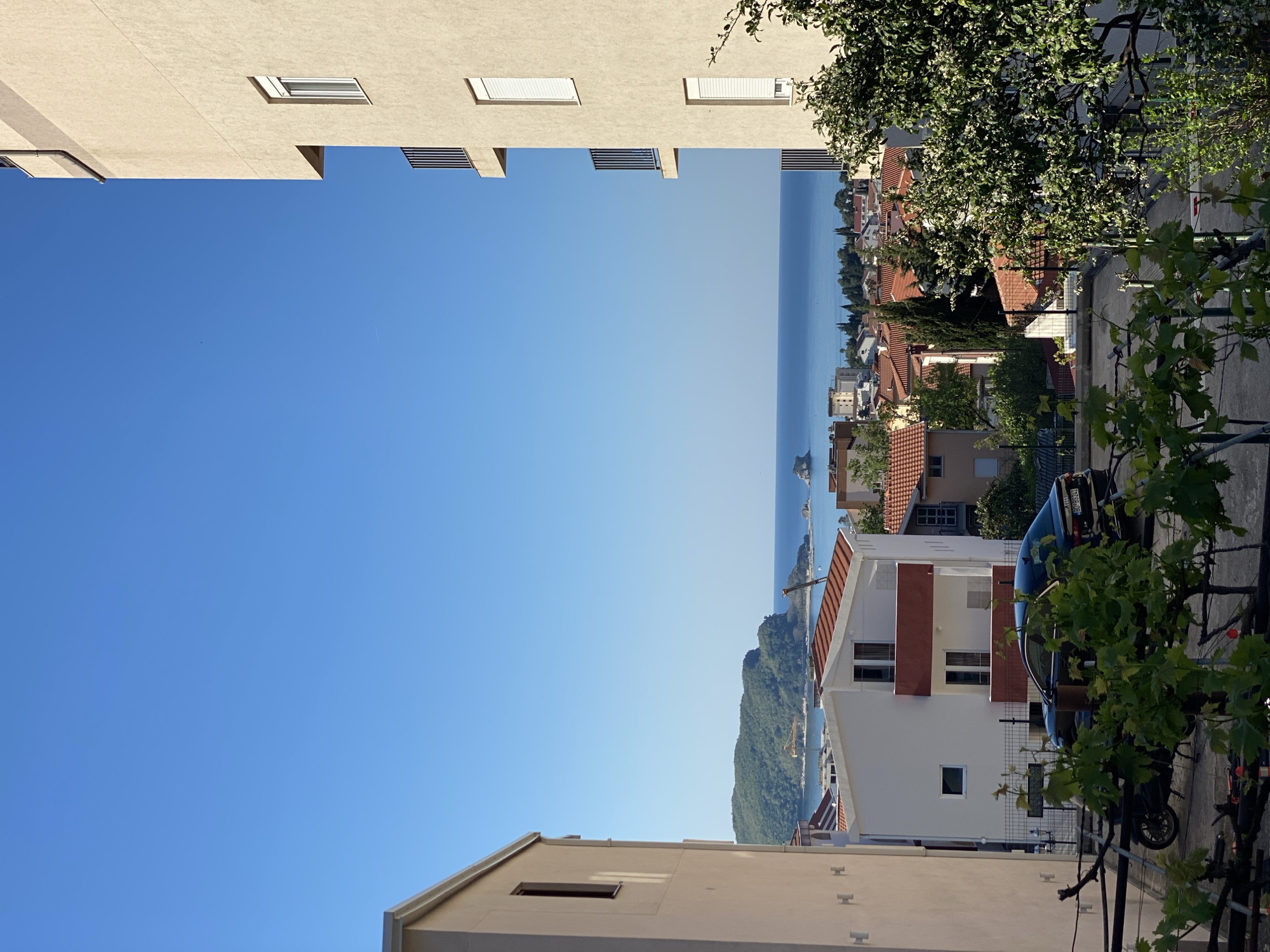 The image shows a view of Budva, Montenegro, framed by buildings on either side. The sky is blue, with a glimpse of the Adriatic Sea in the distance. The right side of the image includes a view of residential buildings with red-tiled roofs, trees, a parked dark blue car, and a vine with green leaves. On the left, the buildings are light-colored. The scene appears to be taken from an elevated position, possibly a balcony or higher floor of a building.