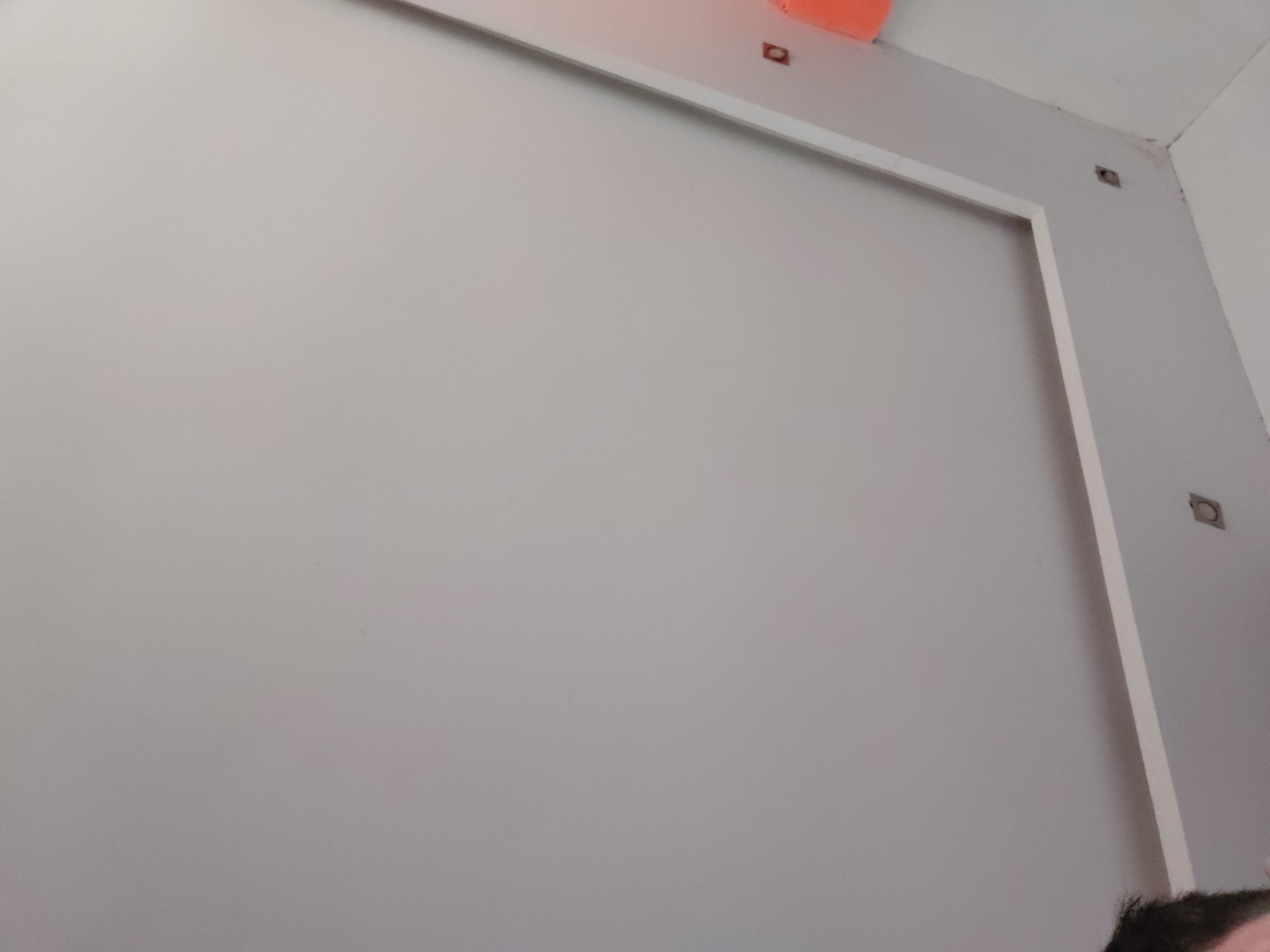 This indoor photograph captures a light gray wall with architectural details, likely taken in Santa Ana, Venezuela. The dominant feature is a large, smooth, light gray wall that occupies most of the frame. A white trim or frame outlines a rectangular section of this wall, suggesting an opening, a door, or a recessed panel.
Above the upper edge of this white trim, a white beam or soffit runs horizontally along the ceiling line. On this beam, two small, dark, square-shaped objects with lighter centers are visible, possibly fasteners, electrical boxes, or mounting hardware. Another similar square object is located in the upper right corner of the image, near the intersection of the wall and ceiling. Partially visible in the top center, resting on or behind the white beam, is a bright orange object, possibly a piece of construction material, tape, or a protective cover.
In the bottom right corner, the top of a person's head, with dark hair, is visible, indicating human presence in the room. The ceiling is white, and the overall impression is of a clean, possibly newly constructed or renovated interior space. There is no visible text in the image. The lighting appears even, suggesting either diffused natural light or artificial illumination.