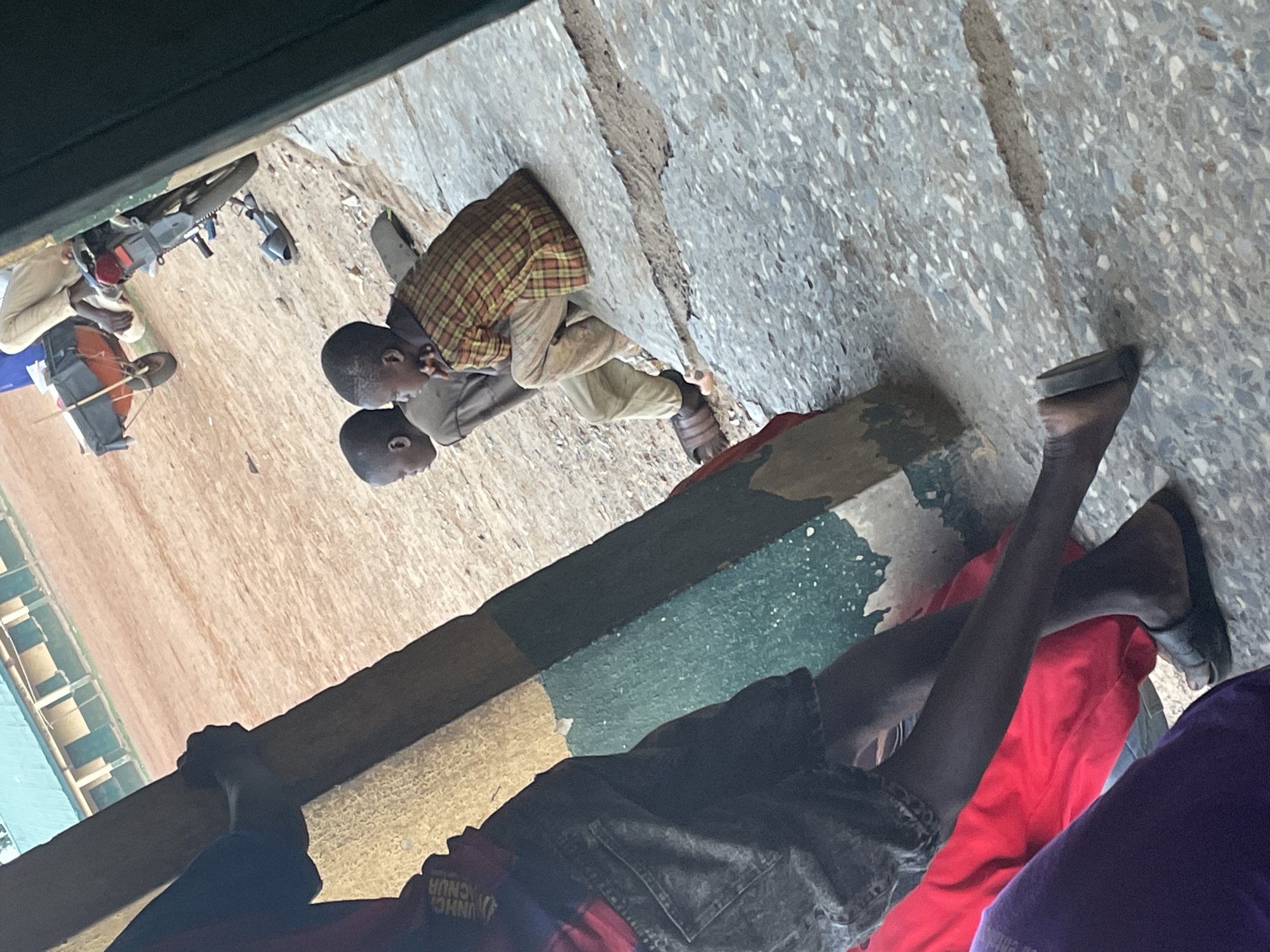 This image, captured in Akwanga, Nigeria, presents an outdoor scene featuring several individuals, primarily children and young adults, in what appears to be a communal or gathering space. The perspective is slightly elevated, looking down upon the subjects.
In the immediate foreground, the lower bodies of three people are visible. One person wears dark denim shorts and a black top, with their dark legs extending into the frame. Another is clad in bright red fabric, possibly trousers, also showing dark legs. A third individual wears purple clothing, partially seen. A dark green, low-lying structure runs horizontally across this section.
Diagonally bisecting the mid-ground is a sturdy, possibly wooden, beam or barrier, painted in alternating sections of dark green, yellow, and blue. Beyond this beam, two young boys are seated or crouching on a light-colored, patterned concrete surface. The boy closer to the viewer is dressed in a light-colored long-sleeved shirt overlaid with a brown and yellow plaid shirt, light trousers, and brown sandals. The other boy, positioned slightly behind him, wears a greyish-brown long-sleeved shirt and light trousers. Both boys have dark skin and short hair, appearing to be resting or engaged in quiet observation.
The background transitions to a reddish-brown dirt or sandy ground. Here, another person is seated near a drum or similar percussion instrument. Further to the left, a dark-colored motorcycle or scooter is parked, with another person nearby. Distant structures with blue-painted sections are faintly visible.
The lighting indicates daytime, with soft, even light suggesting either mid-morning or late afternoon, as there are no harsh shadows. The general atmosphere appears calm and unhurried. A significant detail is the partially visible white text on the red fabric in the foreground, which reads "UNHC" and "ACNUR", identifying a potential connection to the United Nations High Commissioner for Refugees and humanitarian activities. The scene reflects a casual moment of daily life in the Nigerian context.