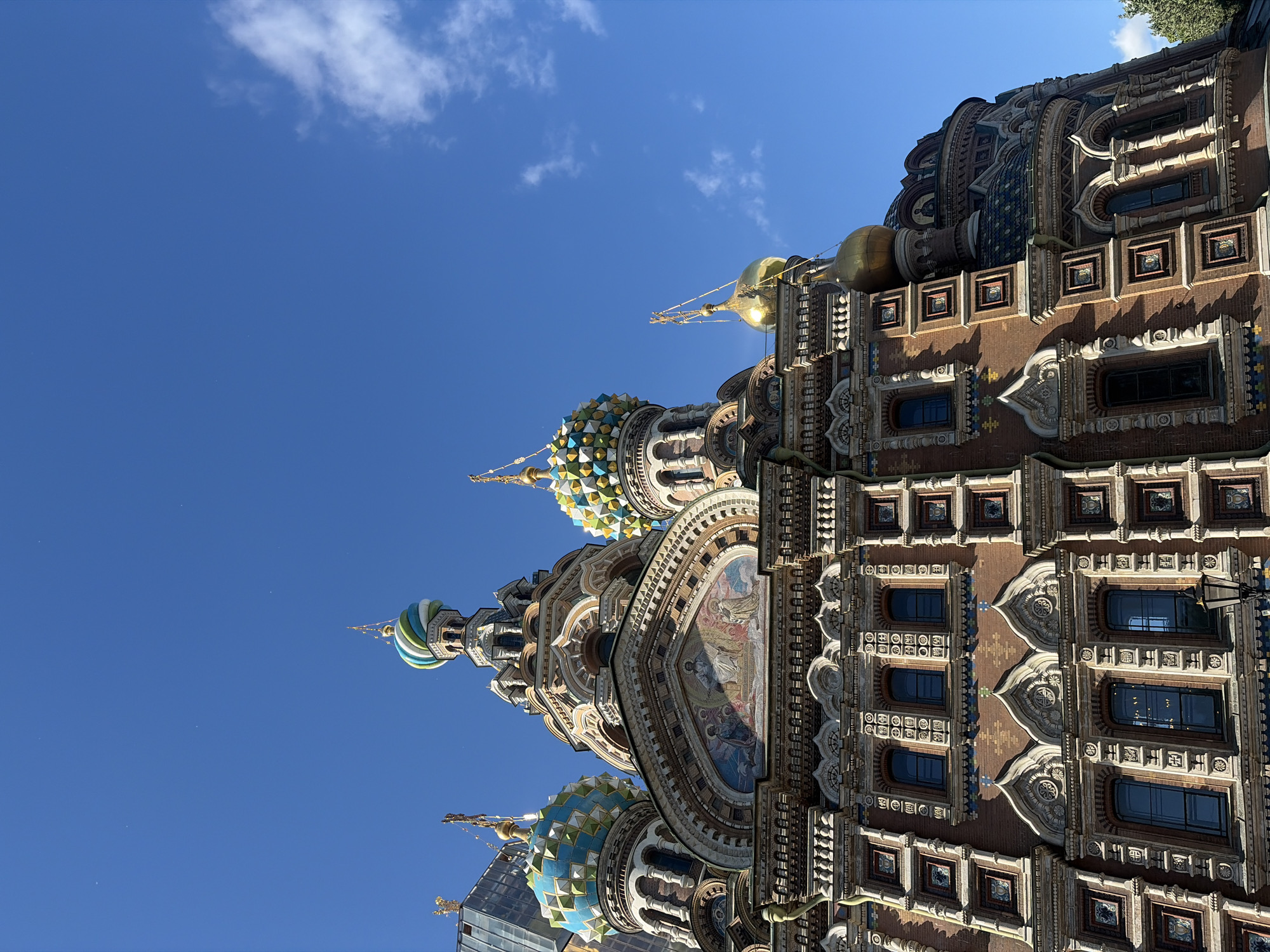 The image captures an upward-angle view of the Church of the Savior on Spilled Blood in Saint Petersburg, Russia. The sky is a clear, vibrant blue with a few wispy white clouds. The church's intricate, multi-colored domes are prominently featured, with one dome adorned with blue, green, and white stripes, and another with a diamond pattern of blue, yellow, and white. The facade of the church is made of red brick with ornate decorative elements, including rows of arched windows, mosaic panels, and golden finials topping the domes and spires. The detailed architecture, typical of Russian Revival style, is evident in the numerous decorative niches and carvings. The lower portion of the image shows a street lamp and the building's textured walls with multiple window openings.