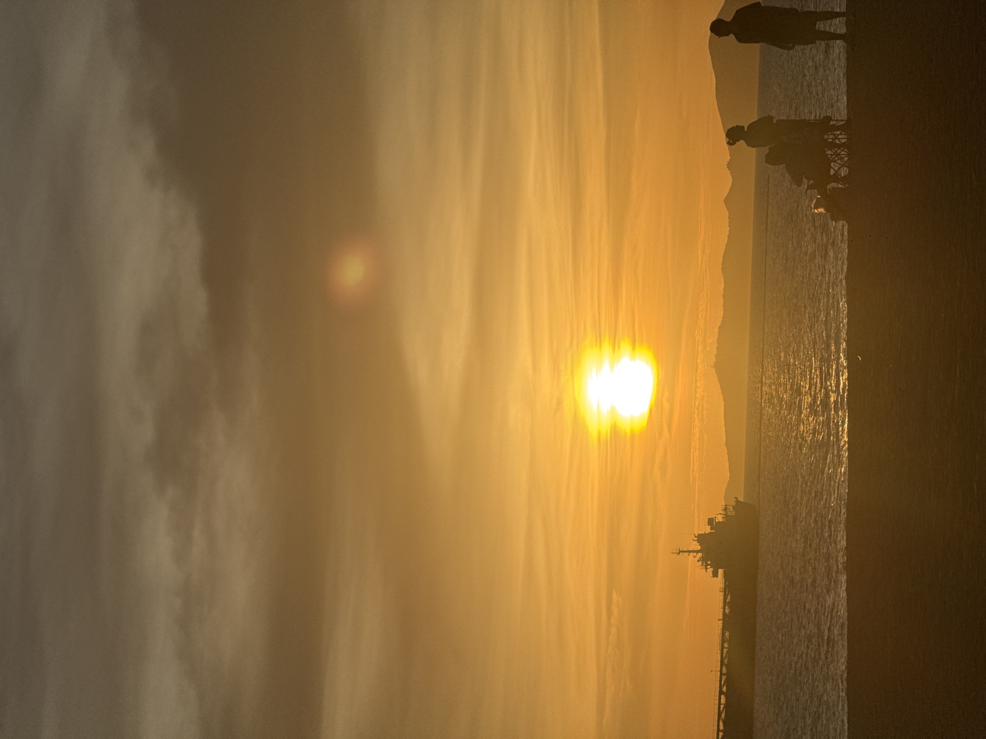 The image captures a vibrant sunset over the ocean in Pattaya City, Thailand. The sun, a bright orb, dominates the horizon. Silhouetted against the radiant sky are figures of people standing on what appears to be a pier or shoreline. The foreground features the silhouette of a cargo ship, adding depth to the scene. The sea reflects the golden hues of the sunset, creating a shimmering effect. The sky is filled with soft, diffused light, indicating a hazy atmosphere. A mountain range appears dimly in the background.