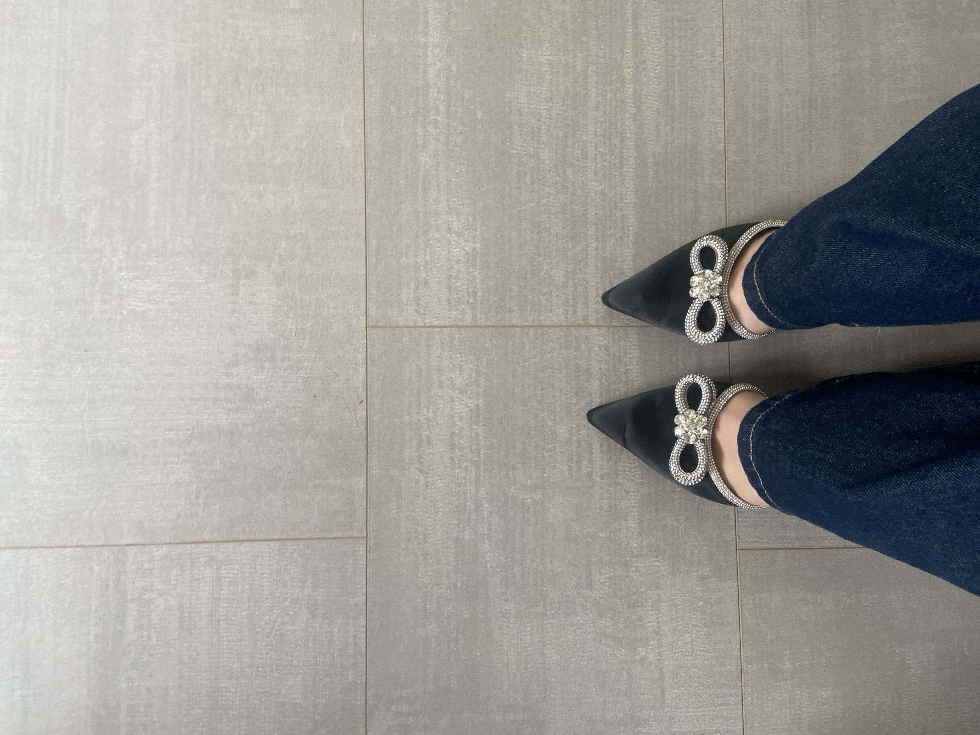 The image shows a pair of feet wearing dark blue jeans and black, pointed-toe dress shoes with a decorative, sparkly bow design at the front of the shoe. The feet are standing on a light grey tiled floor, which has a subtle texture. The tiles are rectangular and arranged in a grid pattern with visible grout lines. The left side of the image is mostly the tiled floor, while the right side features the feet and jeans. The picture is taken from an overhead perspective. There is no visible information to ascertain the location is Dragovishtitsa, Bulgaria.