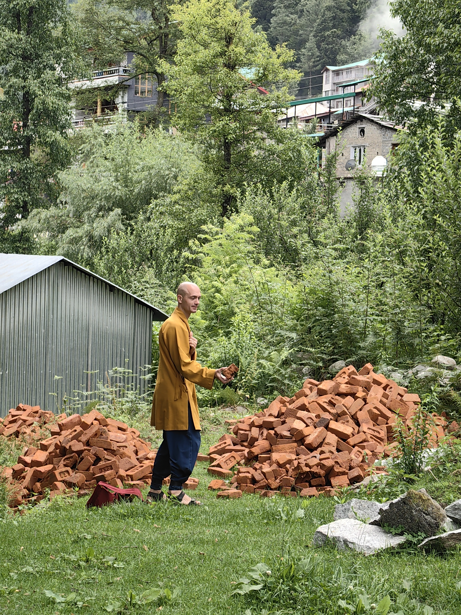 In the city of Vashisht, India, a bald man dressed in a saffron robe and dark pants is standing on a grassy area. He is holding a brick in his right hand, appearing to be in the process of placing it onto a large pile of bricks to his right. To his left, another pile of bricks is visible, along with a maroon bag resting on the grass. Behind the man is a corrugated metal shed. The environment is lush and green, with dense foliage and trees forming a backdrop. In the distance, structures, including buildings with balconies and roofs, are visible nestled among the trees, with some suggesting signs of smoke or steam rising. The overall scene depicts an outdoor setting with construction materials and hints of ongoing activity or life in the surrounding area.