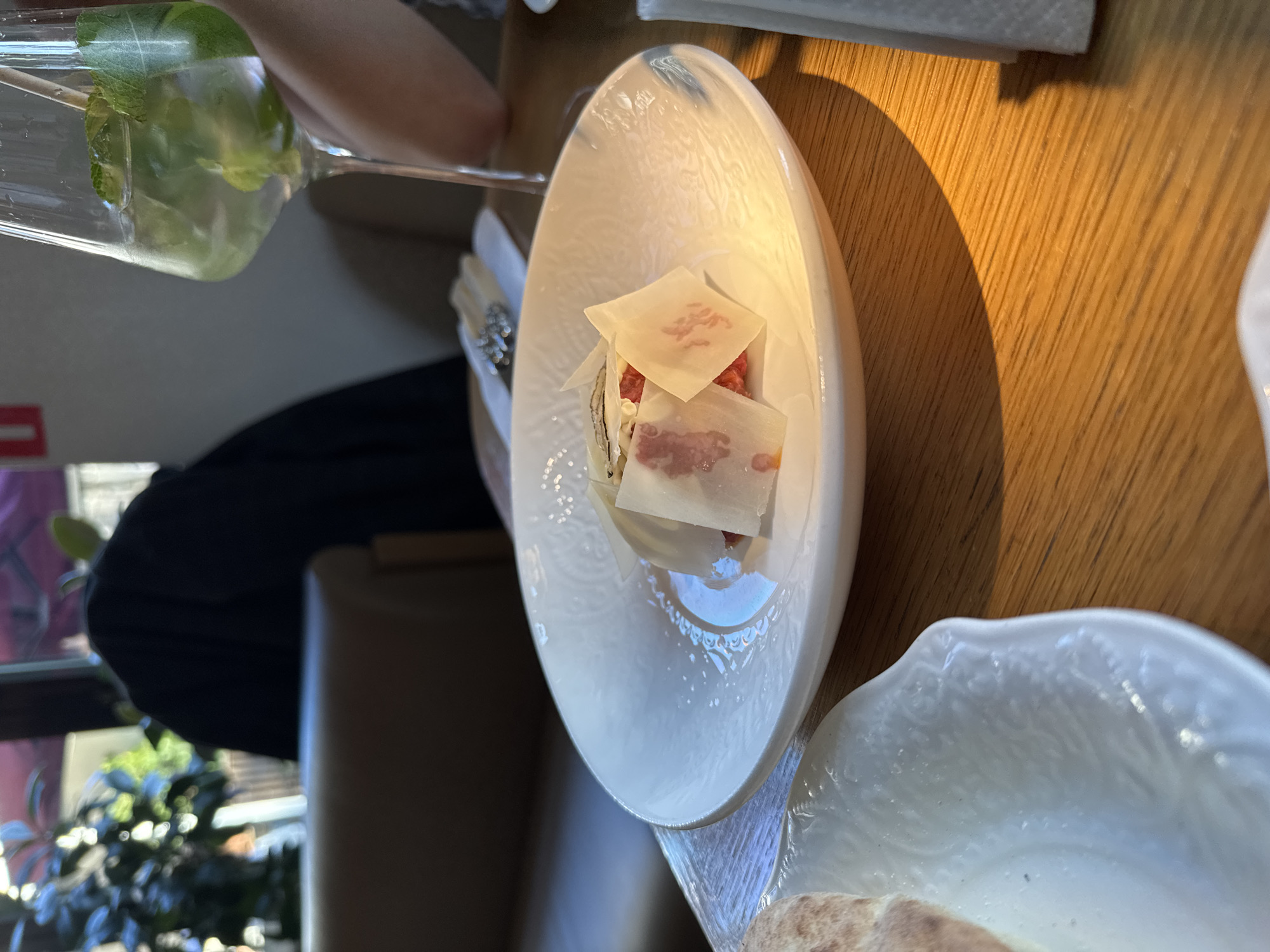This image captures a dining scene in what appears to be a modern restaurant or cafe, likely during the daytime in Moscow, Russia. The shot is taken at a slight angle, focusing on a meticulously presented dish and a drink on a wooden table.
The central subject is a unique white, elongated, and irregularly shaped dish holding a small appetizer. This dish features a base of finely diced red ingredient, possibly a meat tartare or carpaccio, topped with several thin, translucent, rectangular slices. These slices, perhaps cheese or delicate fat, exhibit faint reddish, irregular markings that resemble a pattern or natural marbling rather than legible text.
To the upper left, a hand holds a tall, clear glass containing a light green beverage garnished with fresh mint leaves, possibly a mojito or a similar cocktail, with a straw visible inside. Part of another white dish, possibly containing bread or a pastry, is visible at the bottom right of the frame.
The wooden table surface, with its prominent grain, is bathed in bright, natural light, casting a distinct shadow from an object located out of frame to the left, indicating a clear light source. The background is softly blurred, showing glimpses of an indoor setting with a window overlooking greenery, reinforcing the daytime atmosphere. A person's arm or shoulder, dressed in dark attire, is also partially visible in the blurred background. The scene conveys a sense of calm and elegant dining.
