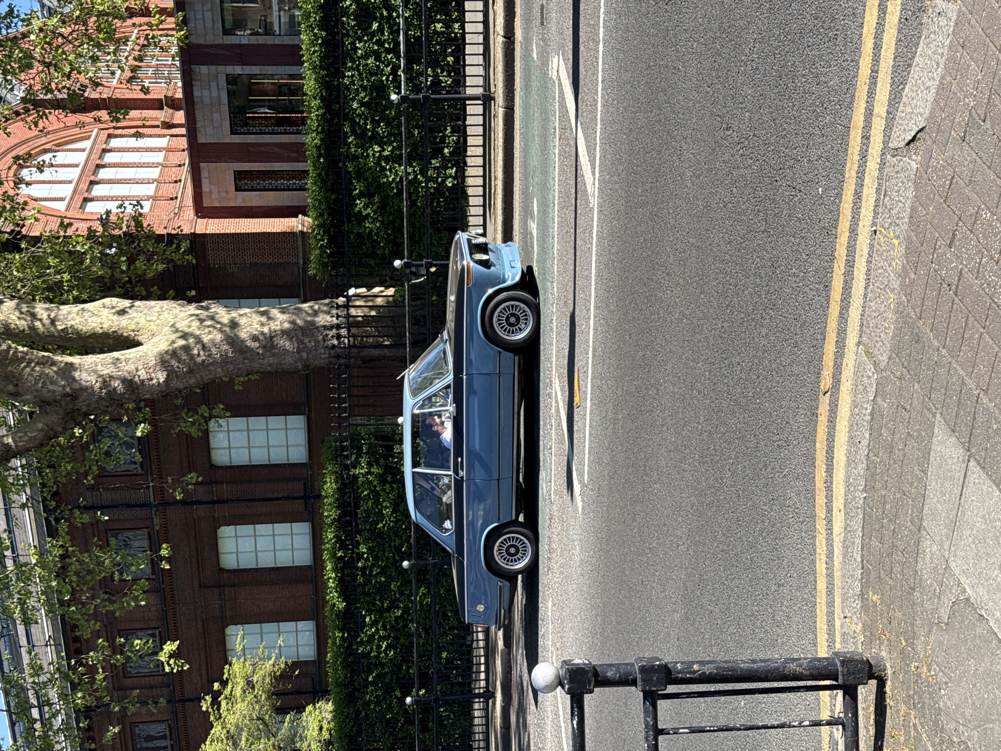 A vintage light blue car, resembling a BMW 2002, is parked on the left side of a paved road under bright sunlight. A person is visible in the driver's seat. The road features double yellow lines near the curb, typical of parking restrictions in the UK. Adjacent to the road, a black metal fence with spherical finials separates the street from a green hedge and trees.
To the left, a substantial tree with a thick, textured trunk and green foliage dominates the midground. Beyond the tree and hedge, a multi-story red brick building with architectural detailing is prominent. The building displays a large arched window with multiple panes on its lower section and various rectangular windows with light-colored frames across its facade. Some windows feature decorative dark grilles.
The scene depicts a sunny daytime setting, with strong shadows cast by the fence and tree, suggesting clear weather. The combination of double yellow lines and the architectural style of the building are strong indicators of a London, United Kingdom location, as provided in the context. No discernible text is visible in the image.