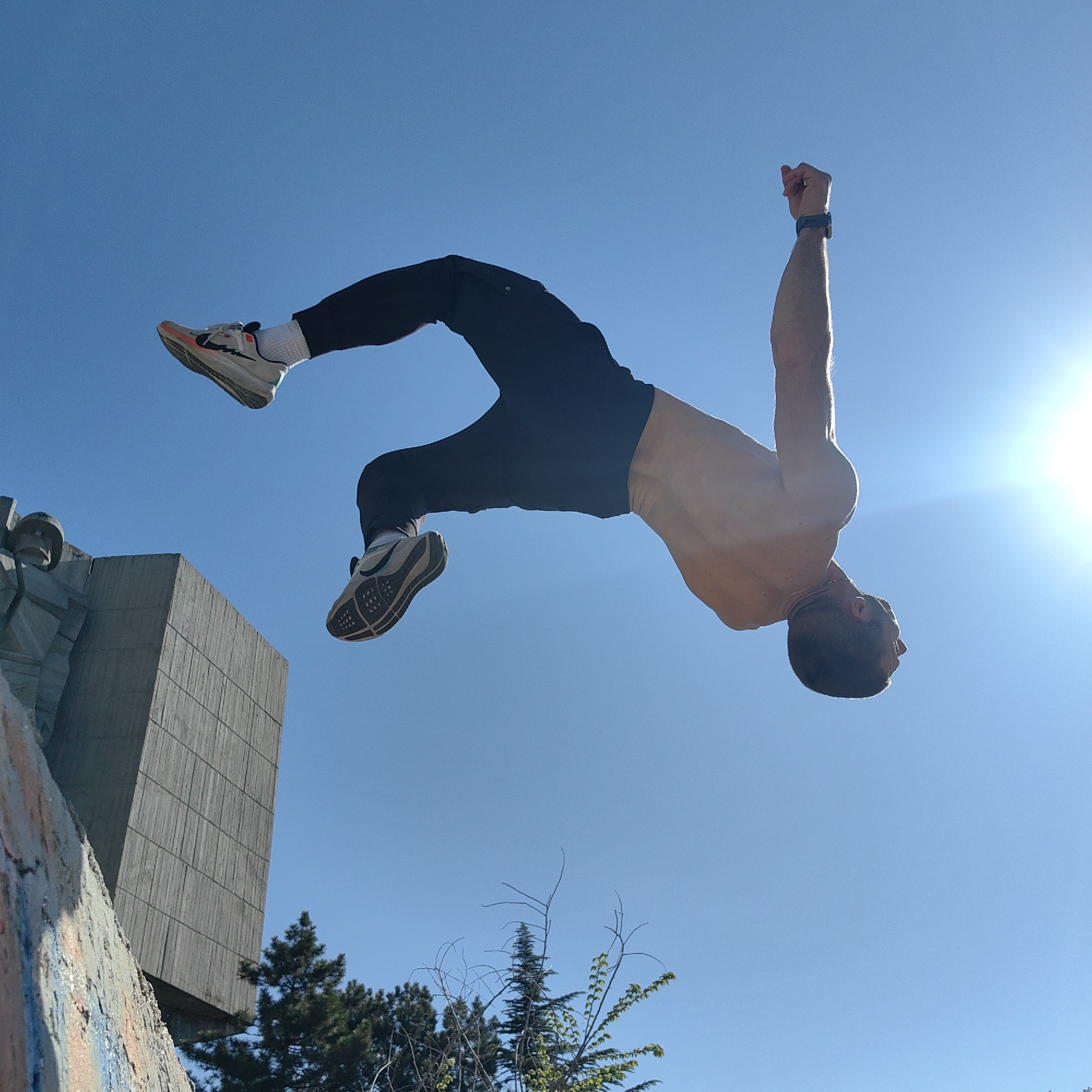The image captures a powerful moment of a shirtless man performing an acrobatic backflip against a vibrant, clear blue sky. He is mid-air and inverted, with his head facing towards the lower right of the frame and his body curled. He wears black athletic pants, white socks, and athletic shoes; one shoe is light-colored with a distinctive red/orange accent and a black swoosh, while the other is beige/grey with a textured sole. A black smartwatch is visible on his left wrist.
The bright sun, positioned in the upper right, creates a strong backlight and lens flare, silhouetting parts of the man's body. In the background, on the left, a large, brutalist-style concrete structure, likely a monument or building, stands tall. It features prominent vertical paneling and includes what appears to be a stylized head or sculptural element. A rugged, possibly graffiti-covered wall occupies the lower left foreground. Green trees and shrubs, along with some bare branches, are visible behind the concrete structure. The scene is brightly illuminated, indicating a clear and sunny day, likely during late morning or early afternoon. No legible text is visible in the image. The setting and architectural style are consistent with locations found in Varna, Bulgaria. The image conveys athleticism and dynamic movement within an urban or monumental environment.