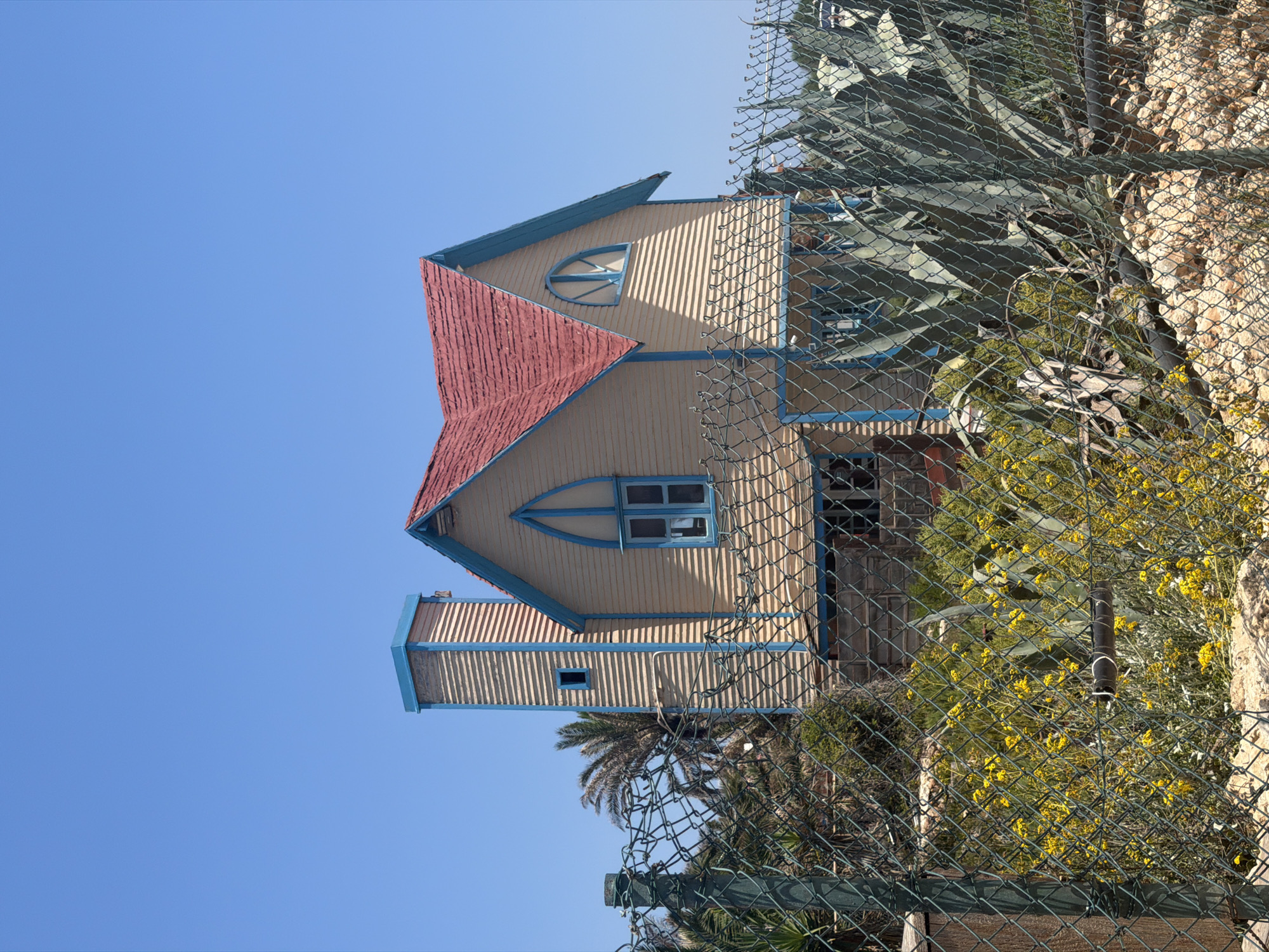 The image shows a whimsical, brightly colored house with a red roof and light yellow siding, accented with blue trim. The house features unique architectural elements, including a distinctive triangular window with blue framing and a smaller rectangular window on a protruding section. The structure is partially obscured by a chain-link fence and lush green foliage, including large agave plants and flowering yellow shrubs. The background is a clear, bright blue sky, suggesting a sunny day. The perspective is from below, looking up at the house, which is tilted on its side in the image. The overall scene evokes a sense of a charming, perhaps slightly surreal, building in a Mediterranean or arid climate. There is no visible text in the image. The location is identified as Mellieħa, Malta.