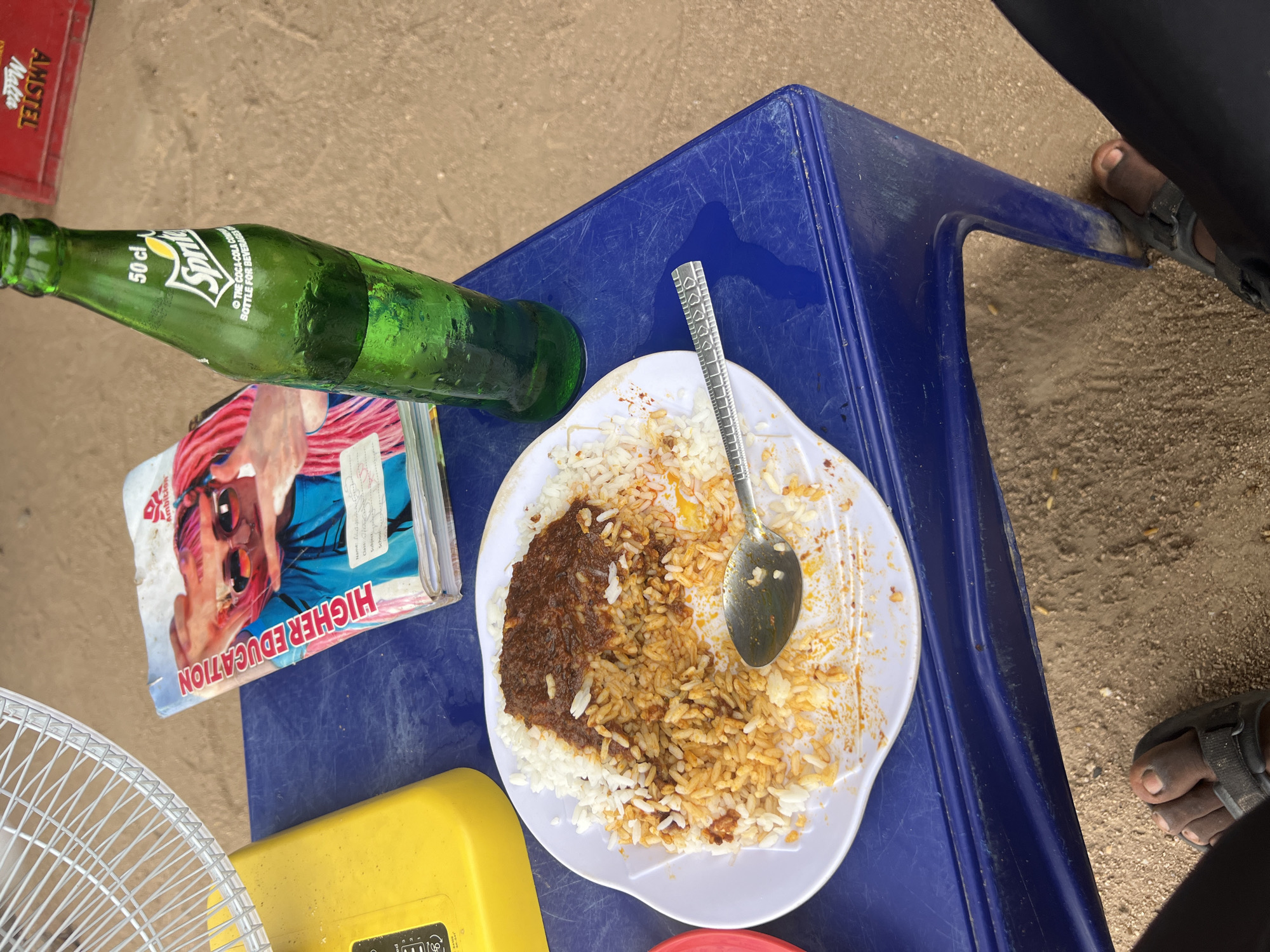 Here's a forensic description of the image:
**Location:** Likely an outdoor setting in Howayi, Nigeria, suggested by the "HIGHER EDUCATION" textbook, "Amstel Malta" beverage, and physical characteristics. The ground appears to be bare earth.
**Subjects:**
* Human: Partially visible are two feet wearing sandals.
* Objects:
* A partially eaten plate of rice and stew with a spoon resting on the food.
* A textbook titled "HIGHER EDUCATION."
* A 50 cl Sprite glass bottle.
* A yellow object of unknown type, partially visible.
* A blue plastic table.
* The edge of a fan.
* A red Amstel Malta case.
**Actions:** Eating (incomplete).
**Interactions:** Human interaction with food and objects on the table.
**Environment:** Bright outdoor lighting. The overall setting appears casual and likely public.
